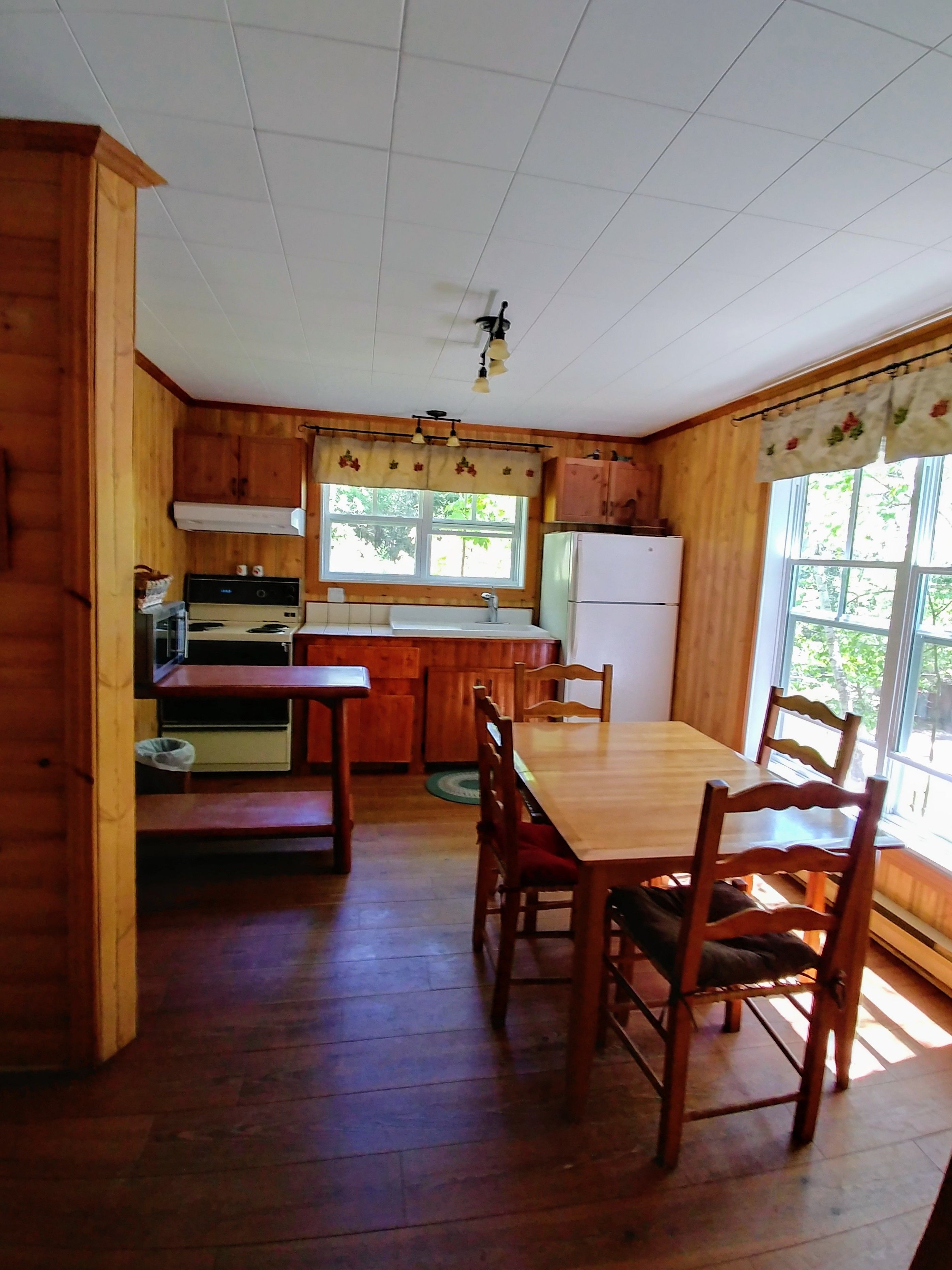 Een houten keukentje in chaletstijl met een eettafel, stoelen, een koelkast en een raam met gordijnen.