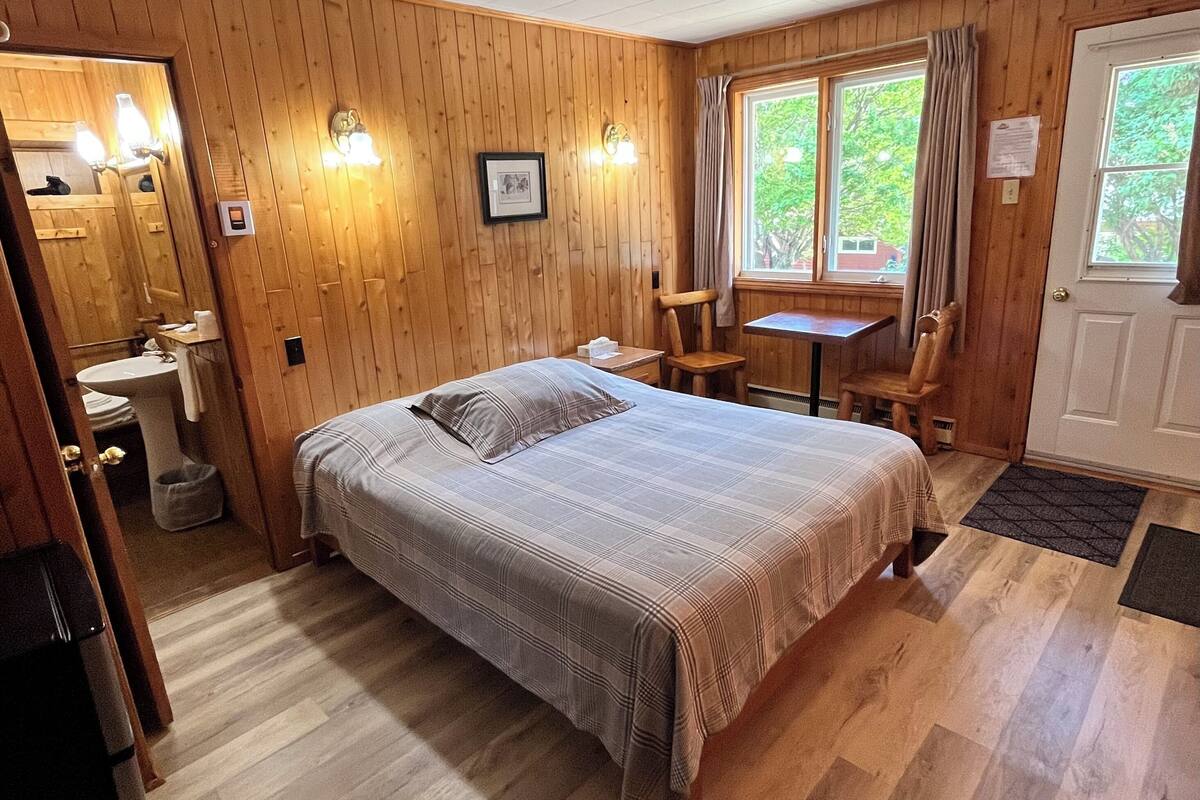 Et rum med træpaneler, seng, vask, spejl og et vindue med gardiner.