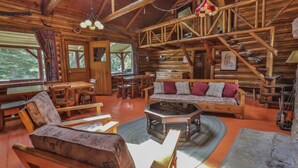 A rustic cabin interior with wooden furniture, a sofa with cushions, a dining table, and a staircase. - Kan-à-Mouche Pourvoirie Auberge et Chalets (Saint-Michel-des-Saints)