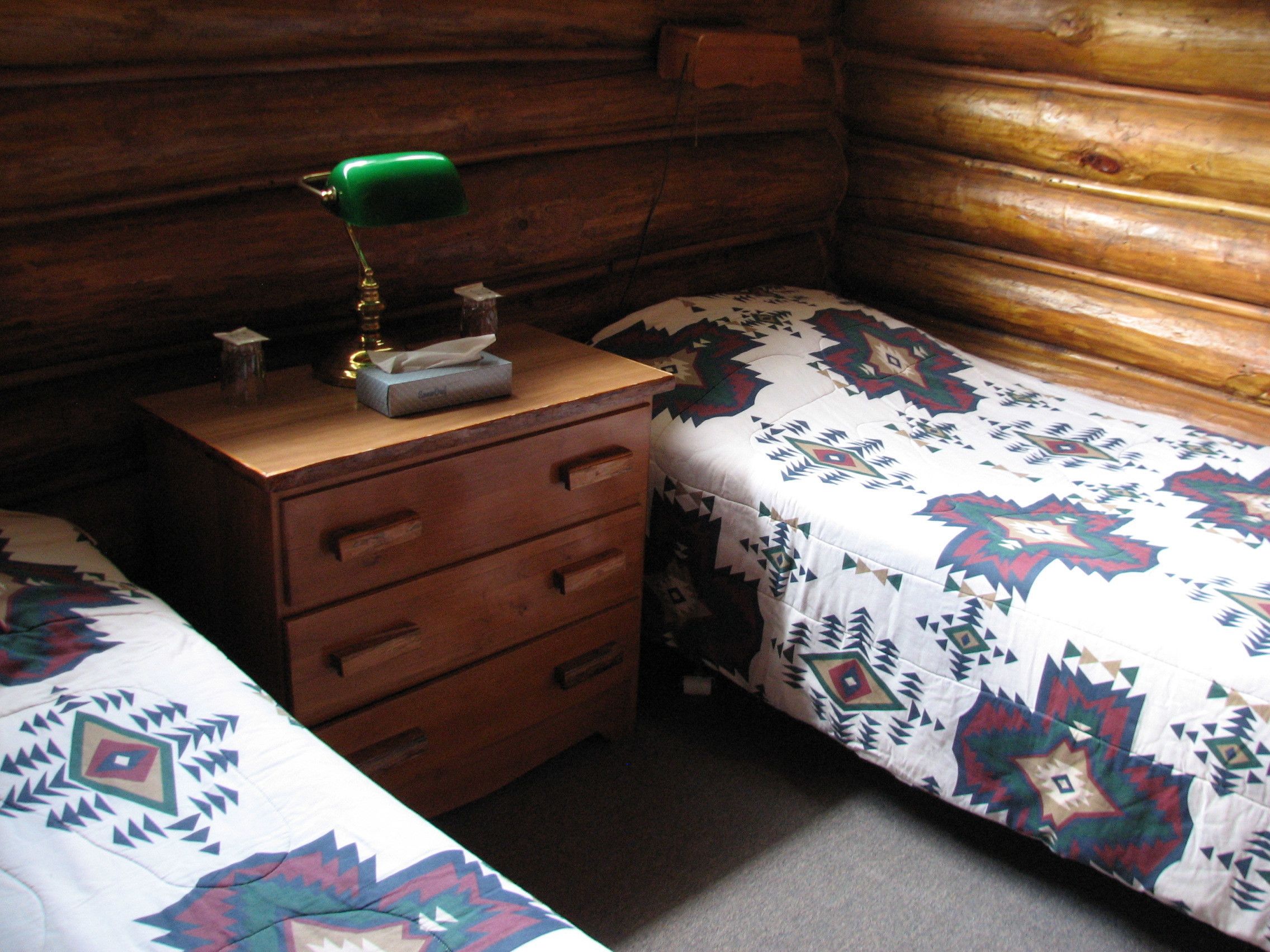 A small, cozy room with wooden walls and a single bed with a patterned bedspread. There is a wooden nightstand with a green lamp and a tissue box on it.