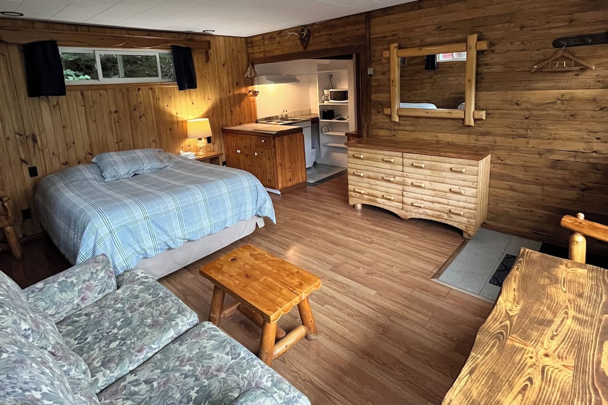 Et hyggeligt værelse i sommerhusstil med en seng, en sofa, en trækommode og et lille bord.