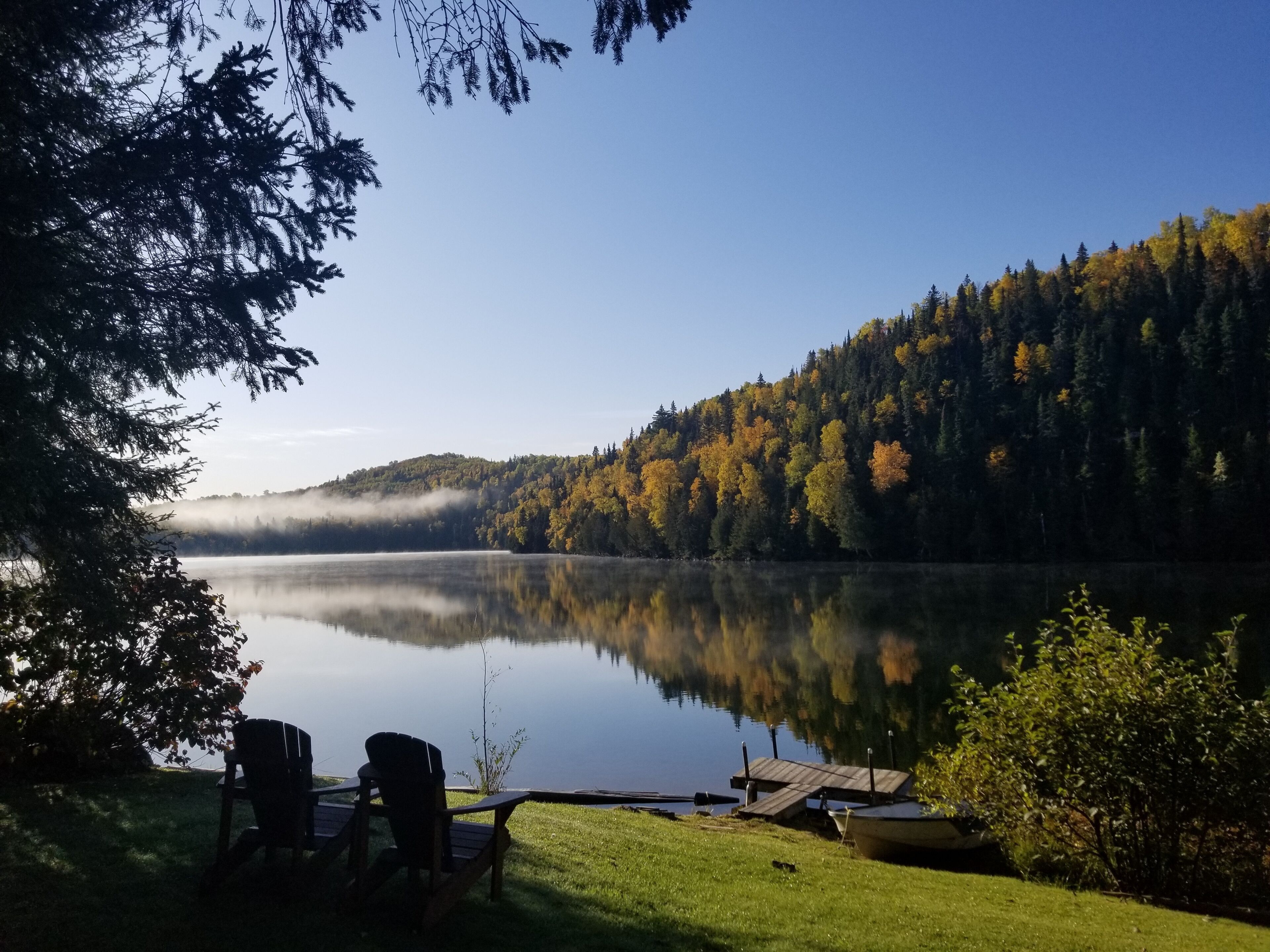A serene lakeside view with a dock, chairs, and a boat, surrounded by autumn-colored trees and a misty hill.