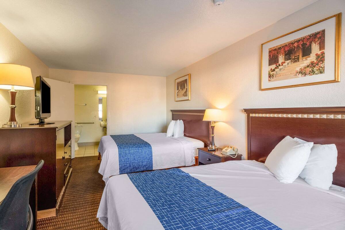 A hotel room with two beds, a desk, a chair, a TV, and a bathroom visible through an open door.