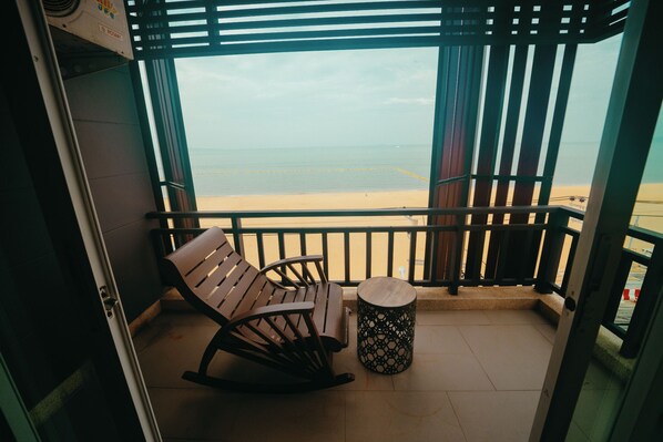 A balcony with a wooden rocking chair and a round table, overlooking a sandy beach and the sea.