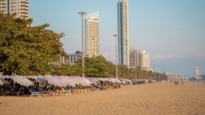 A beach with rows of beach chairs and umbrellas, tall buildings in the background, and palm trees. - Seaside Jomtien Beach (Pattaya)