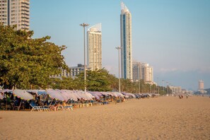 Une plage bordée de rangées de chaises longues et de parasols, avec de grands immeubles en arrière-plan et des palmiers.