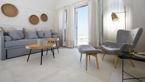 A modern living room with a sofa, round coffee table, and armchair, a floor lamp, and a view through a large window. - De Sol Spa Hotel (Santorini)