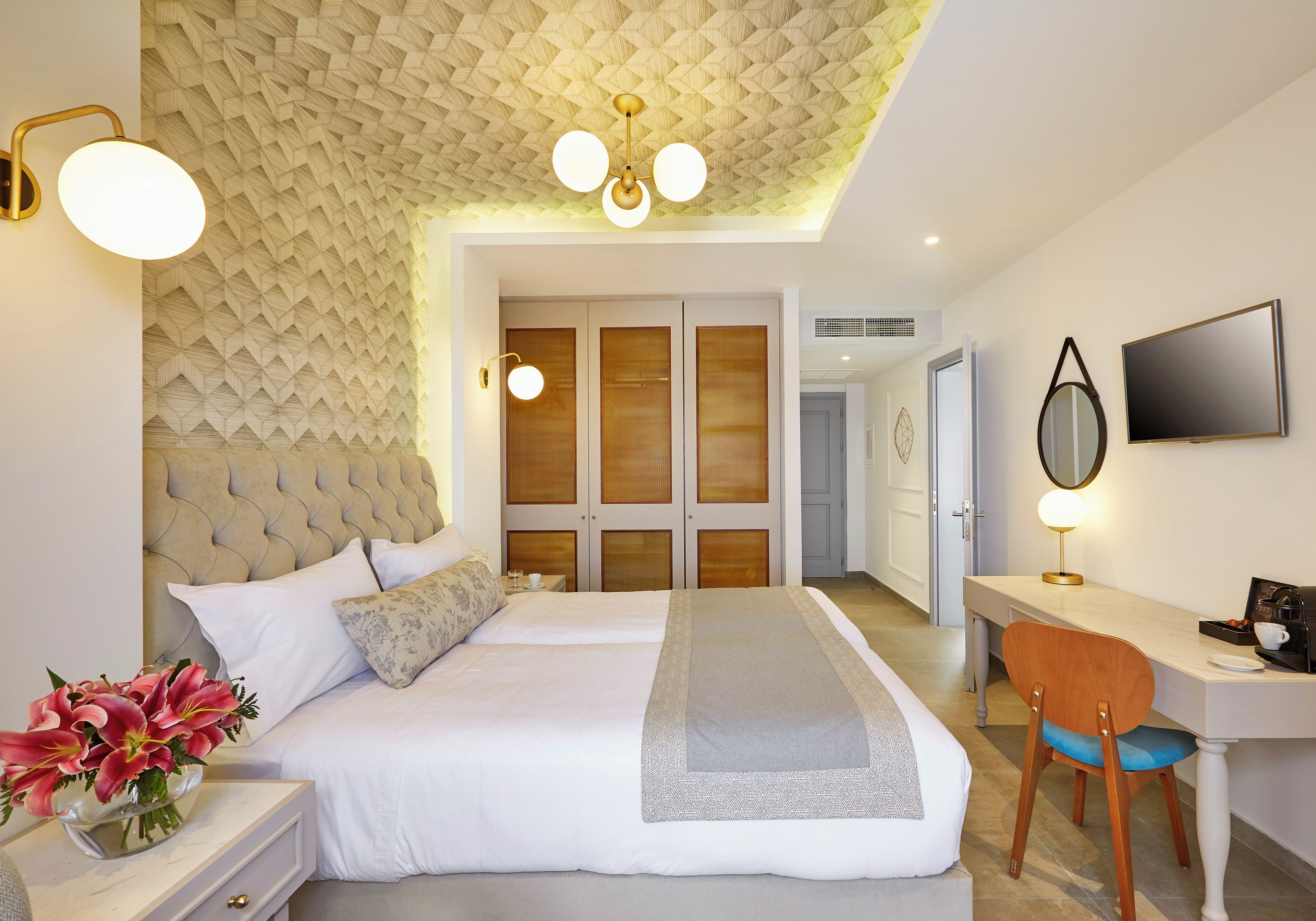Une chambre moderne avec une tête de lit capitonnée, un grand lit recouvert de linge de maison blanc, un bureau avec une chaise et un téléviseur fixé au mur.
