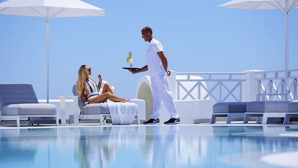 A poolside area with lounge chairs and umbrellas, a waiter serving a drink to a seated woman. - De Sol Spa Hotel (Santorini)