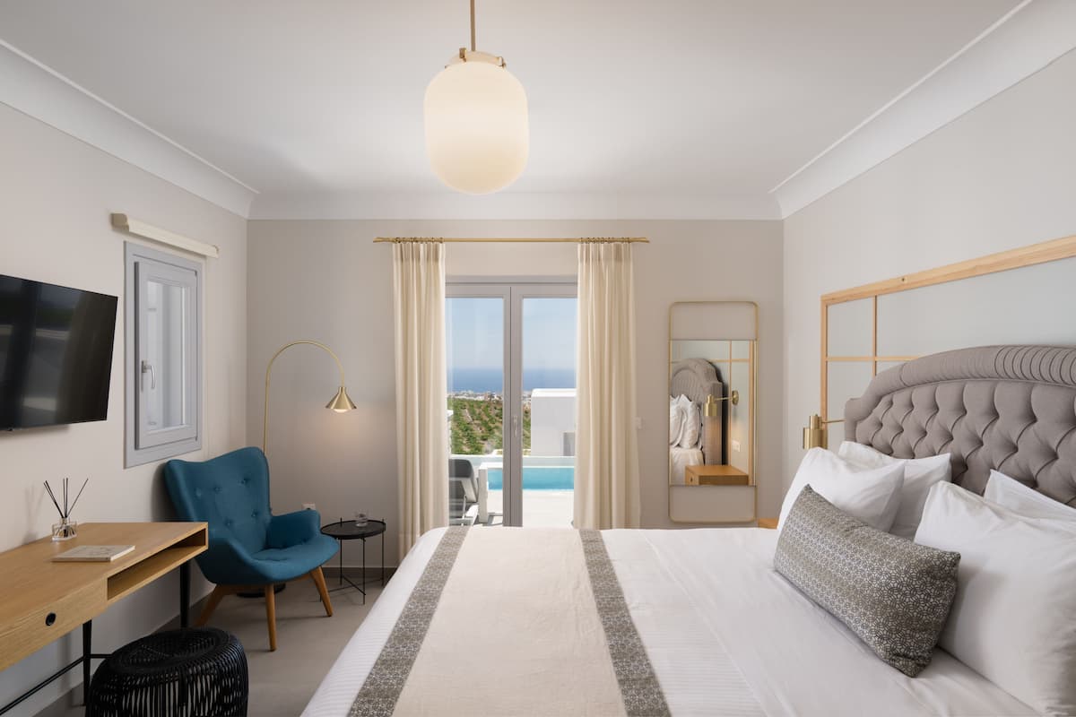 A modern hotel room with a large bed, a desk, a blue armchair, a TV, and a view of the sea through the balcony doors.
