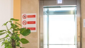 An elevator with a glass door, a potted plant, and a sign with red and white symbols on the wall. - Hotel Camposol (Benidorm)