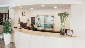 A reception desk with a clock, a framed certificate, and a large potted plant. - Hotel Camposol (Benidorm)