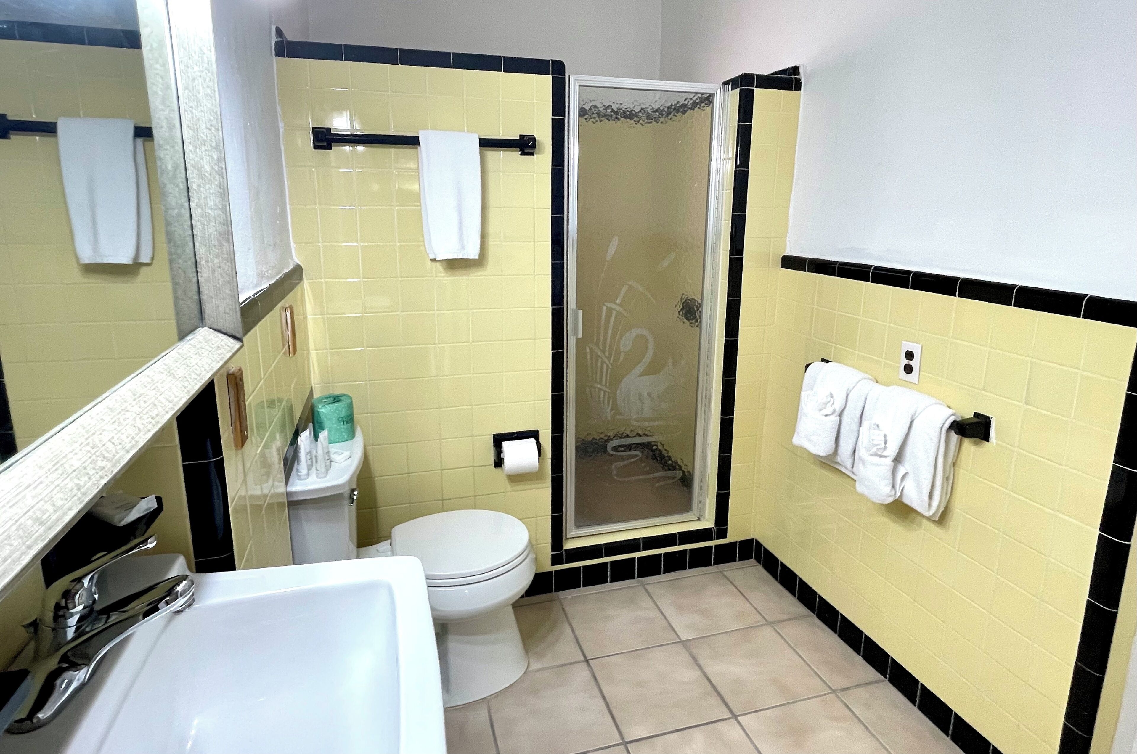 Un baño con paredes revestidas de azulejos amarillos y negros, un lavamanos blanco, un inodoro y una ducha con mampara de vidrio.