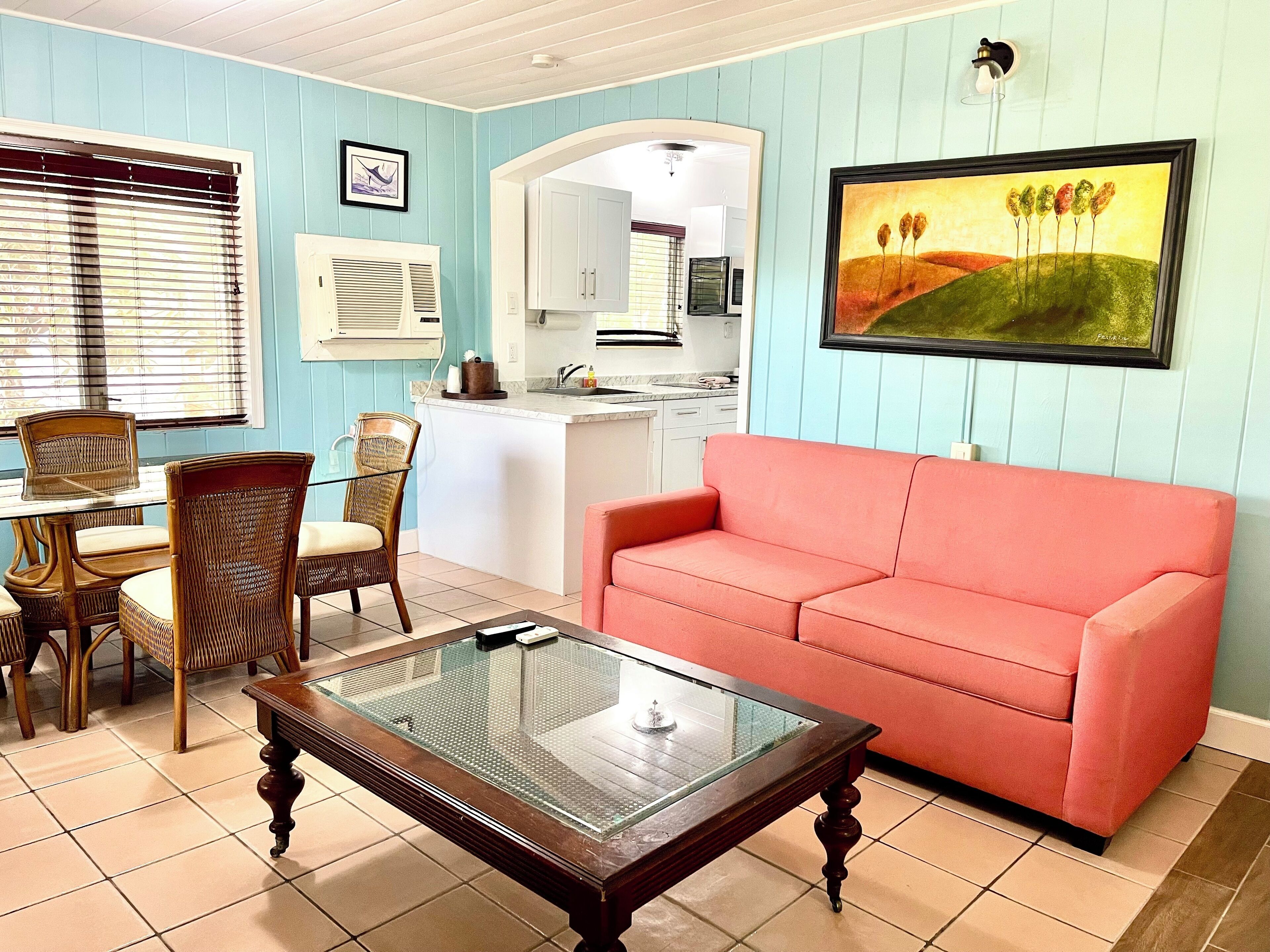 A living room with a glass-top coffee table, a red sofa, wicker chairs, and a dining table with chairs.