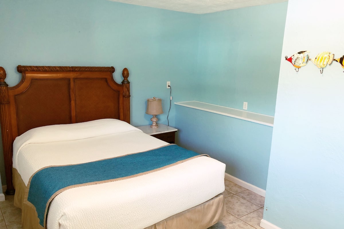 Une chambre à coucher avec une tête de lit en bois, un lit recouvert d’une couvre-lit blanche et bleue, une table de chevet avec une lampe et une étagère murale agrémentée d’objets décoratifs.