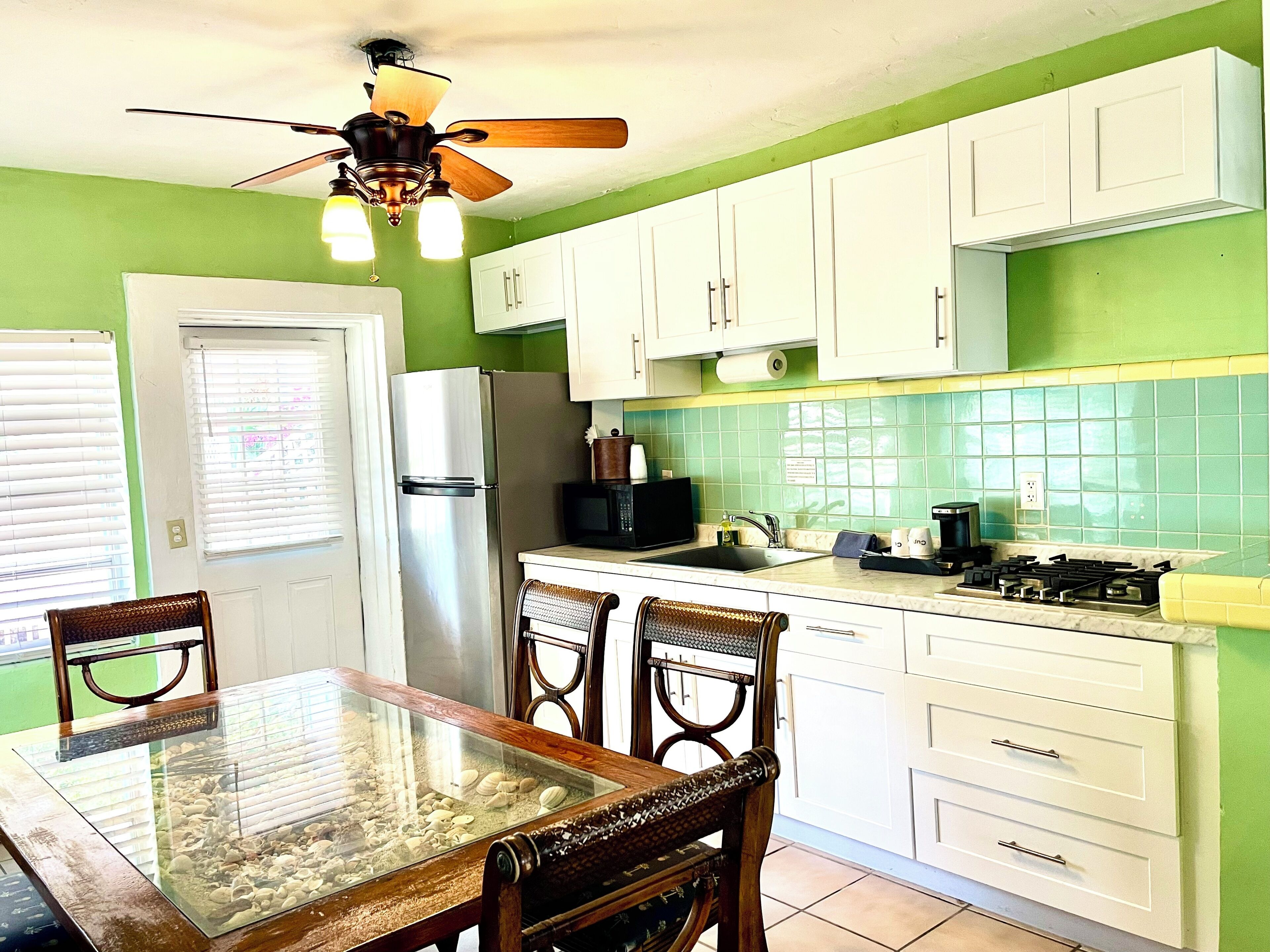 A kitchen with white cabinets, a stainless steel refrigerator, a glass-top dining table, and a ceiling fan.