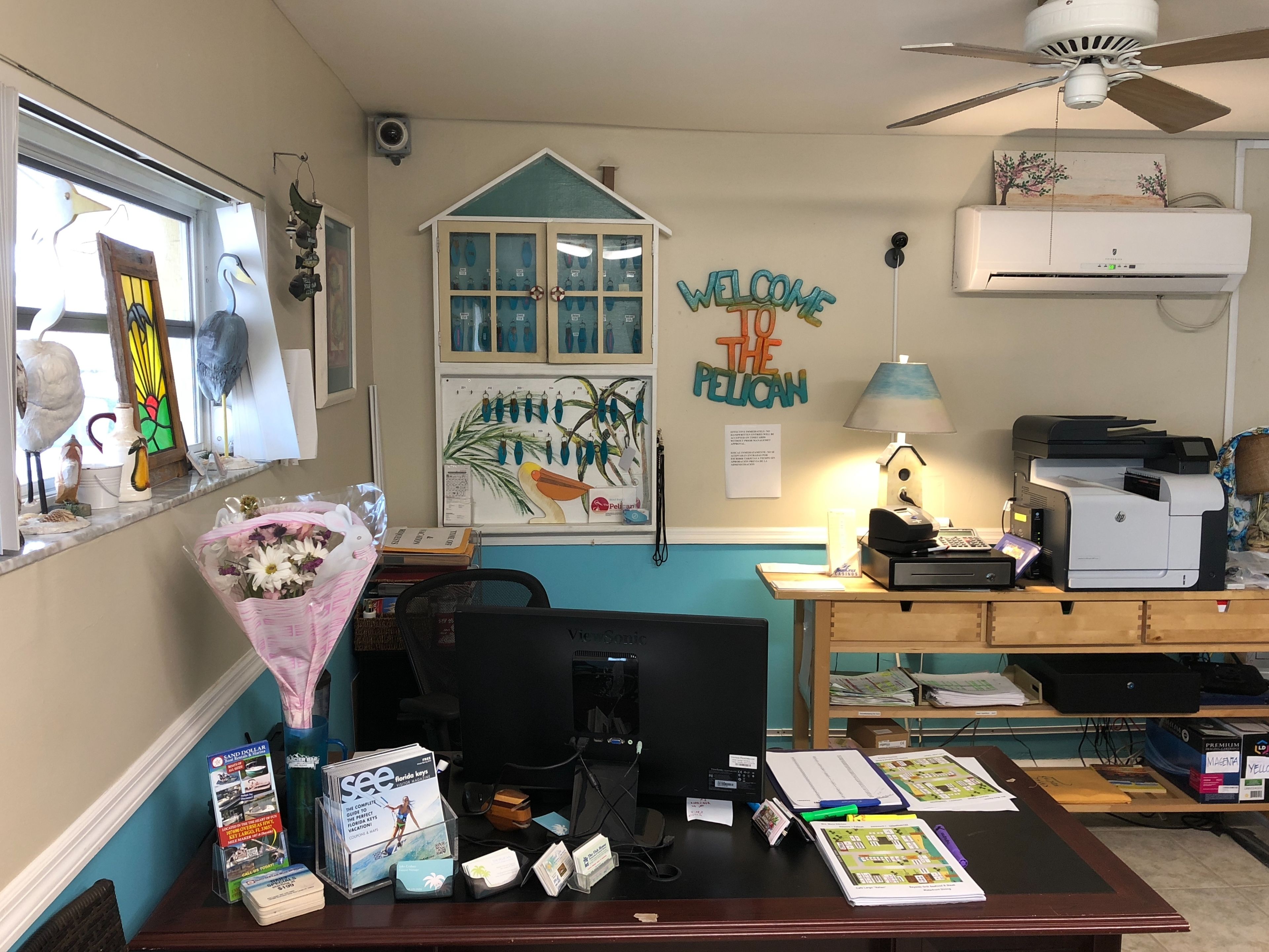 Taulell de recepció amb un ordinador, una impressora i diferents material d'oficina. Hi ha una finestra amb objectes decoratius, un aire condicionat fixat a la paret i un cartell que diu 'Welcome to the Pelican'.
