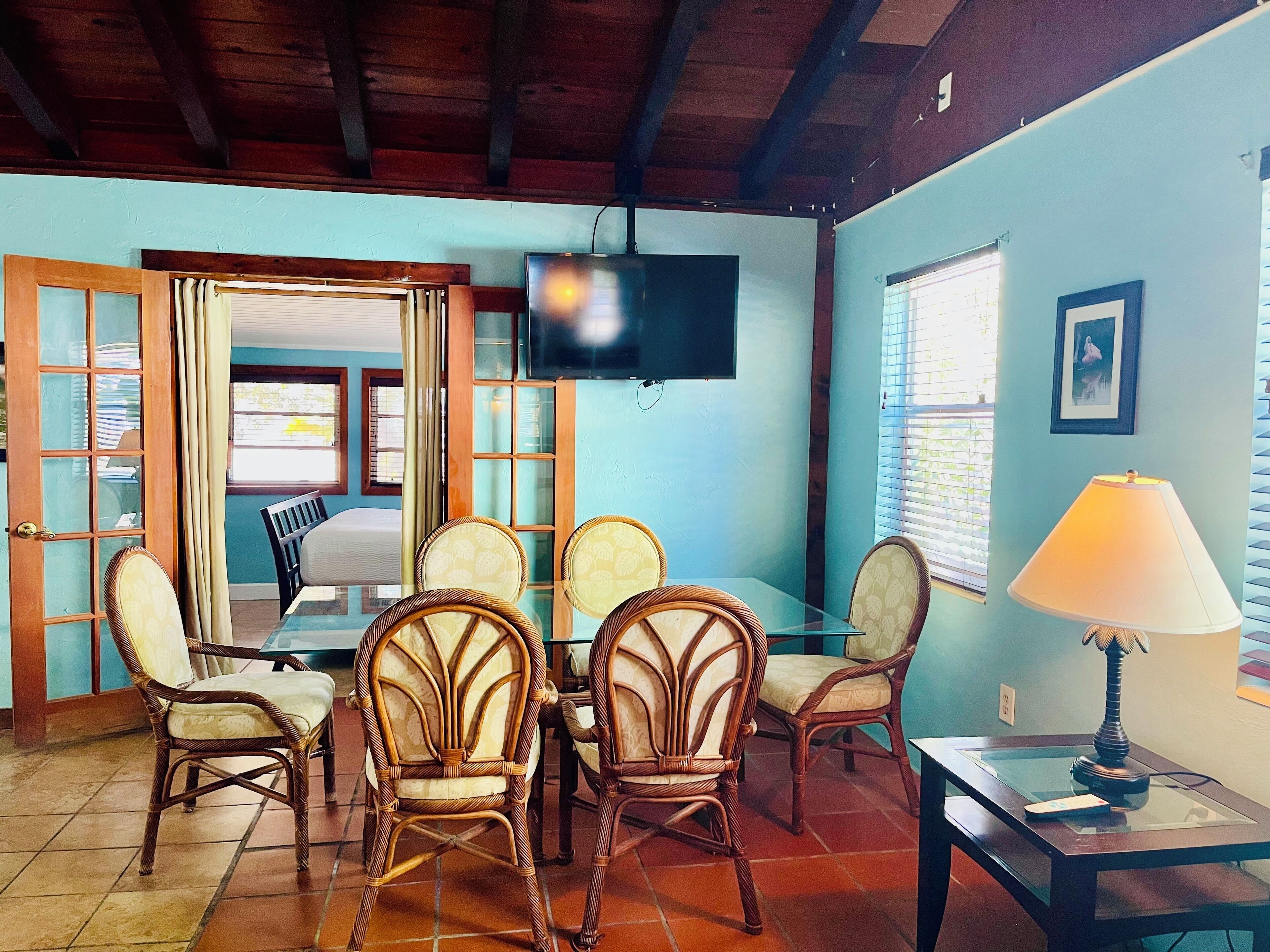 A room with a dining table, four wicker chairs, a TV mounted on the wall, and a lamp on a side table.