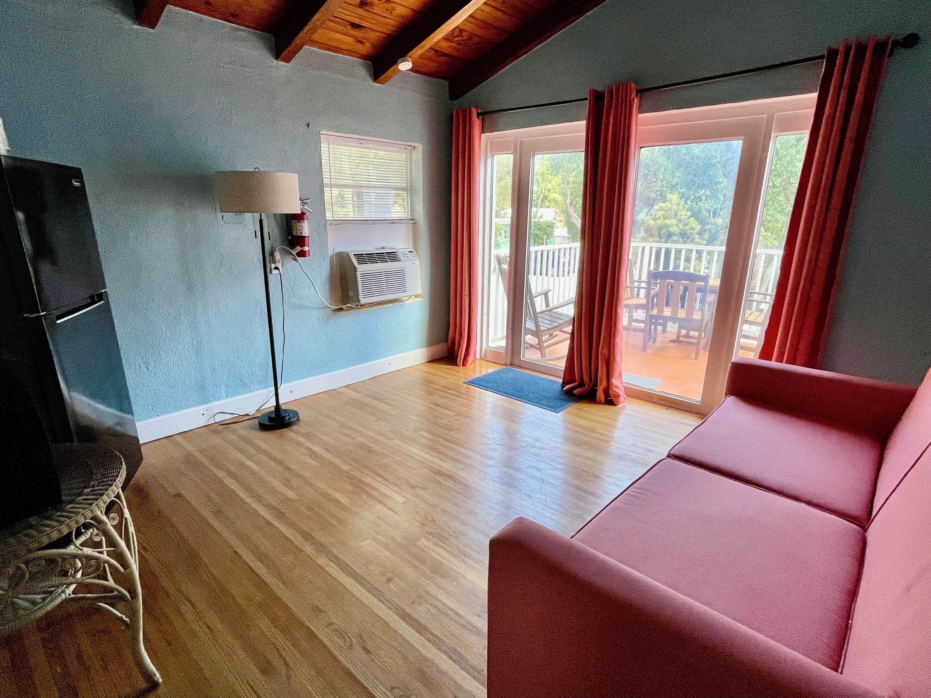 A room with a wooden floor, a red sofa, a lamp, a ceiling fan, and a sliding glass door leading to an outdoor area with a patio.