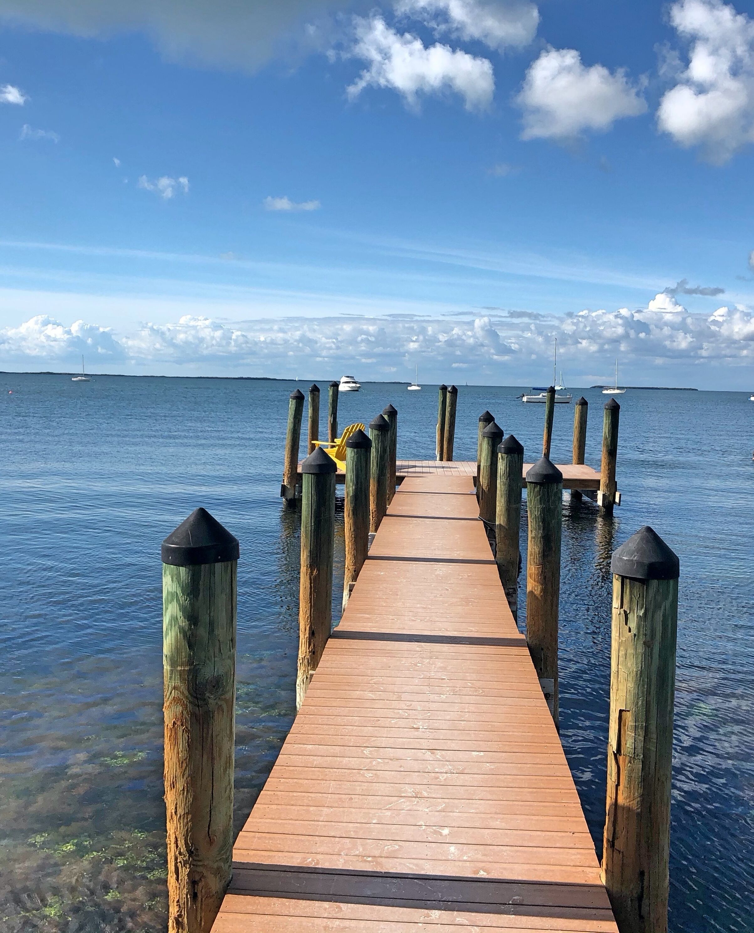 A wooden pier extending into calm waters, with boats anchored in the distance and a clear sky above.