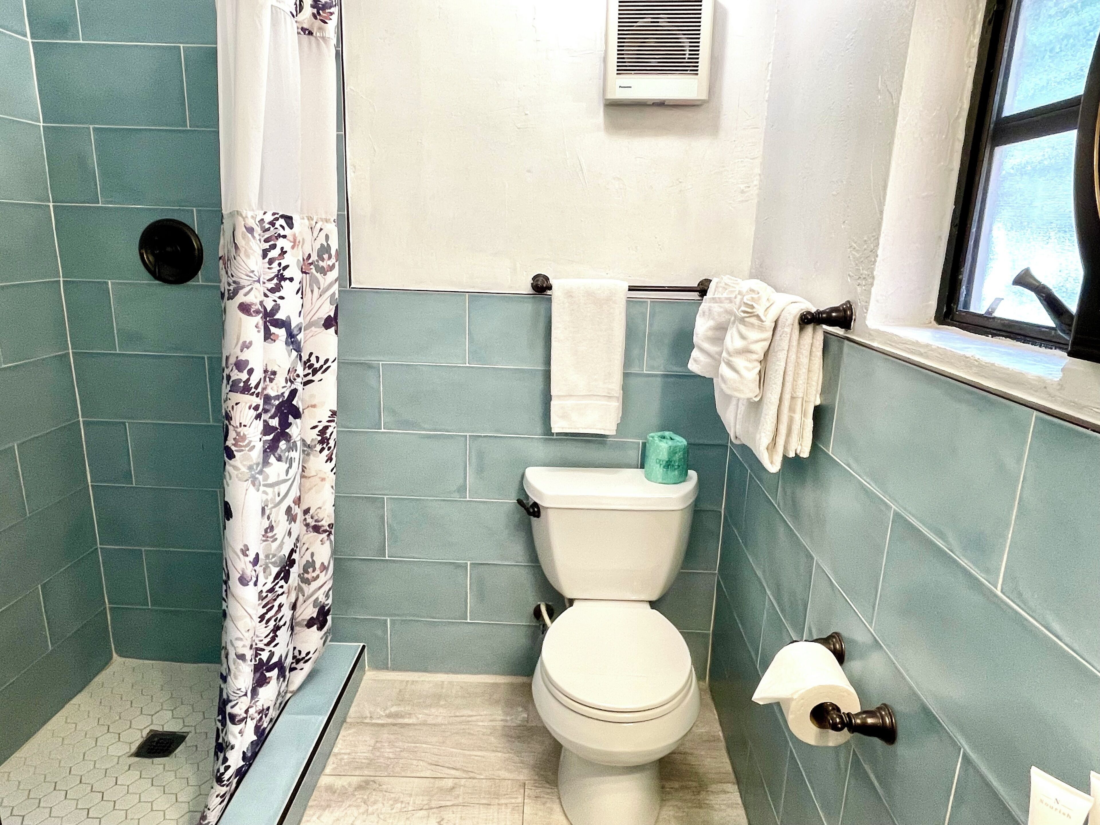 Een badkamer met smaragdgroene tegels, een toilet, een douchegordijn en een raam.