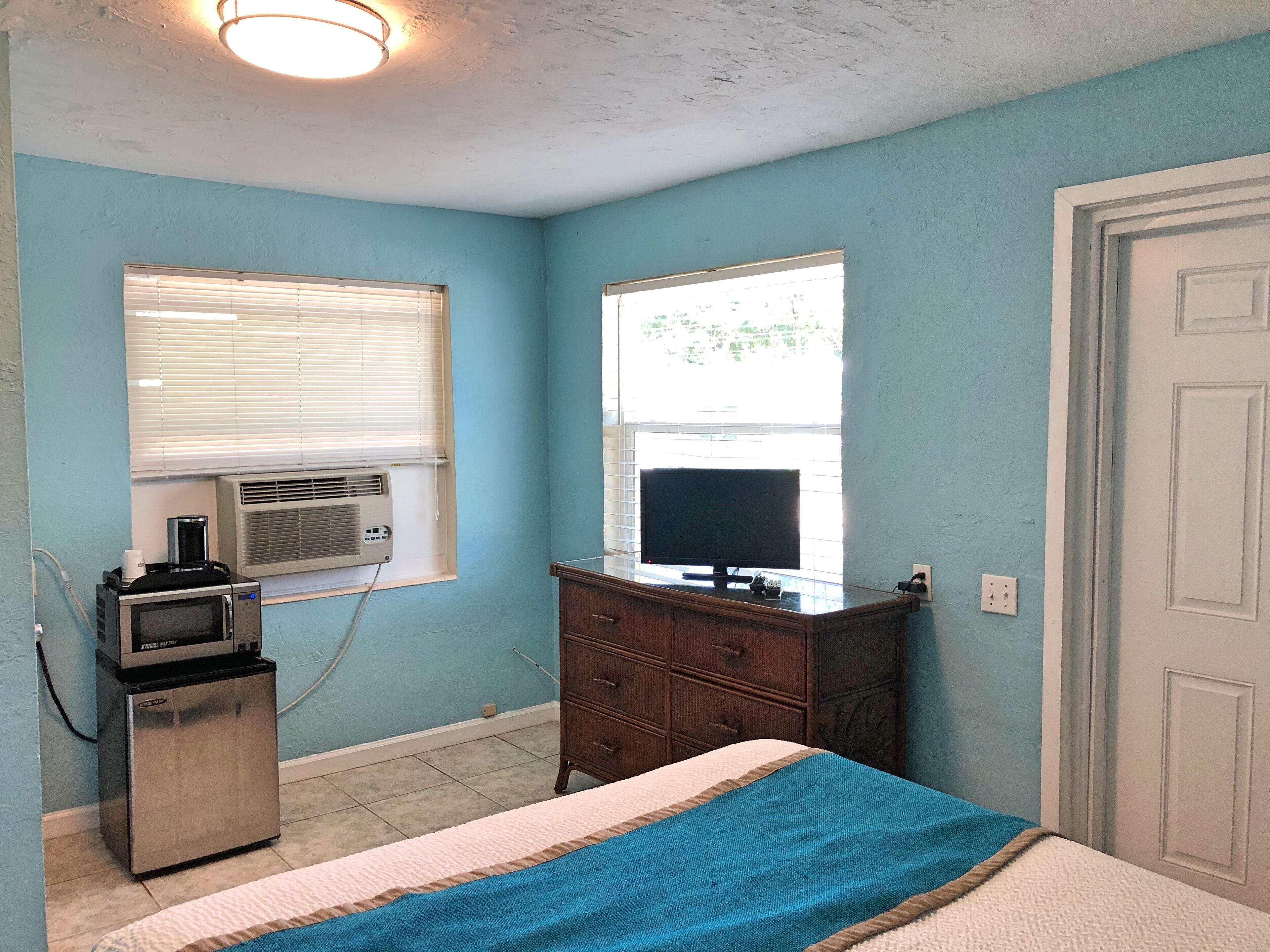 A bedroom with a bed, a dresser, a television on a stand, an air conditioner, and a window with blinds.