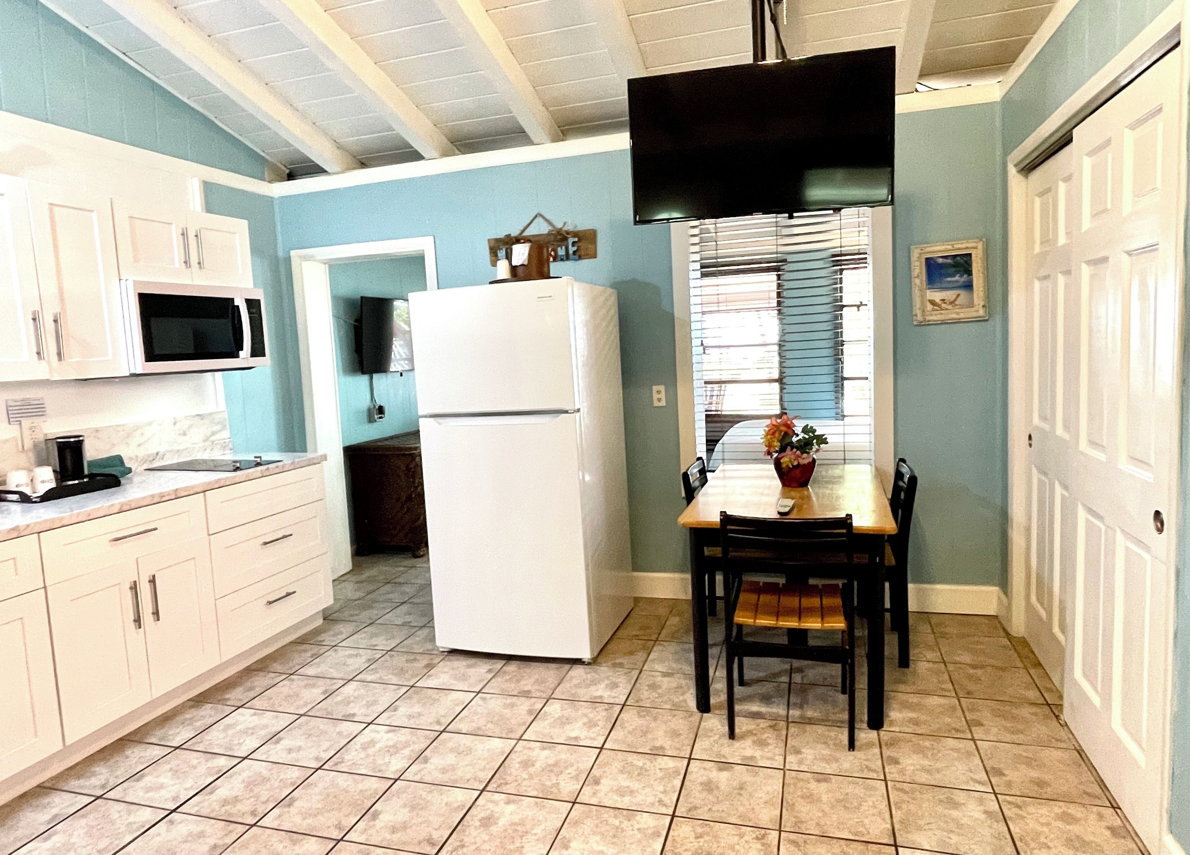 Una cocina con gabinetes blancos, un refrigerador blanco, una mesa de comedor con sillas y un televisor montado en la pared.