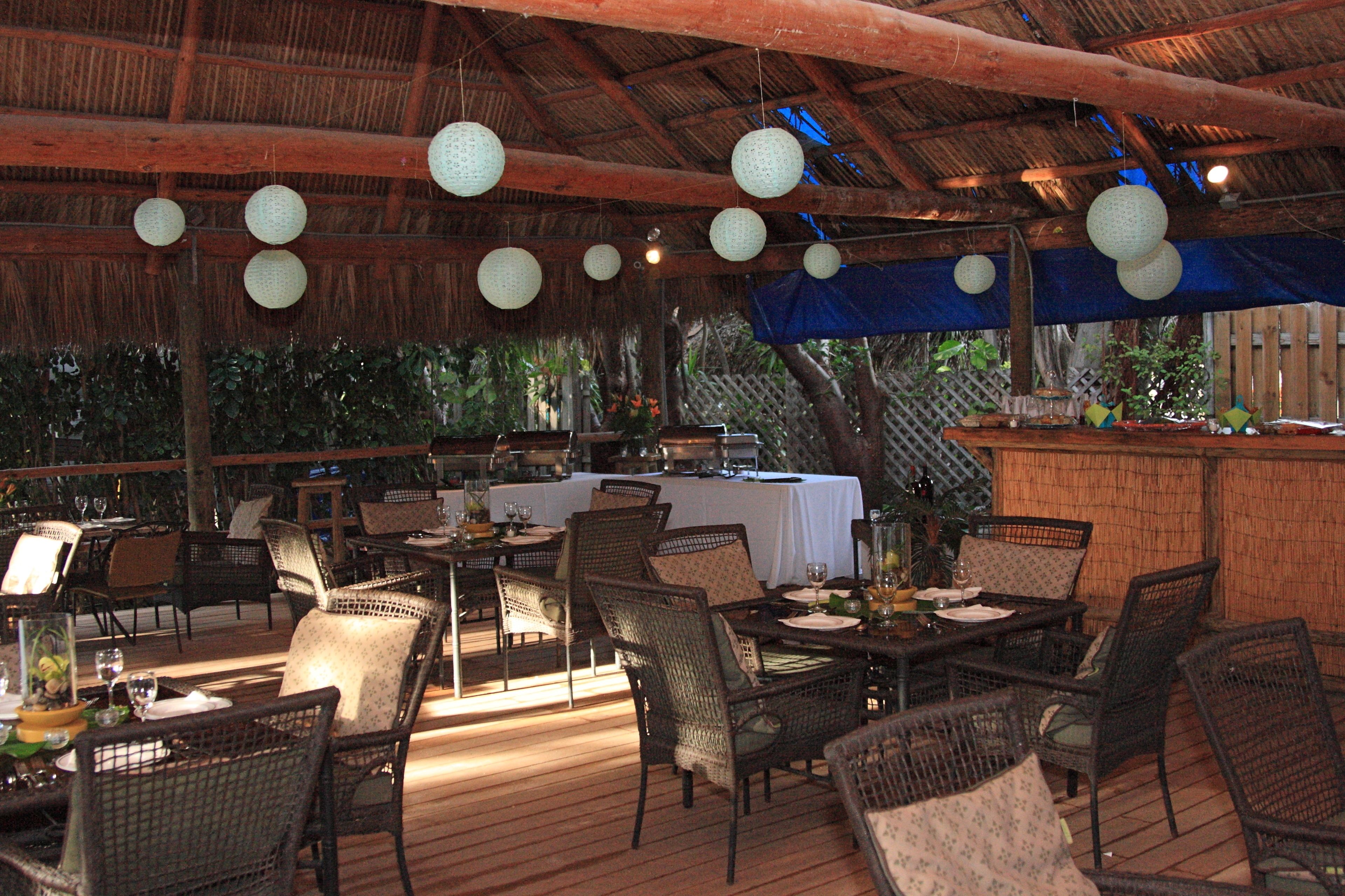 An outdoor dining area with wicker furniture, tables set with plates and glasses, and hanging lanterns.
