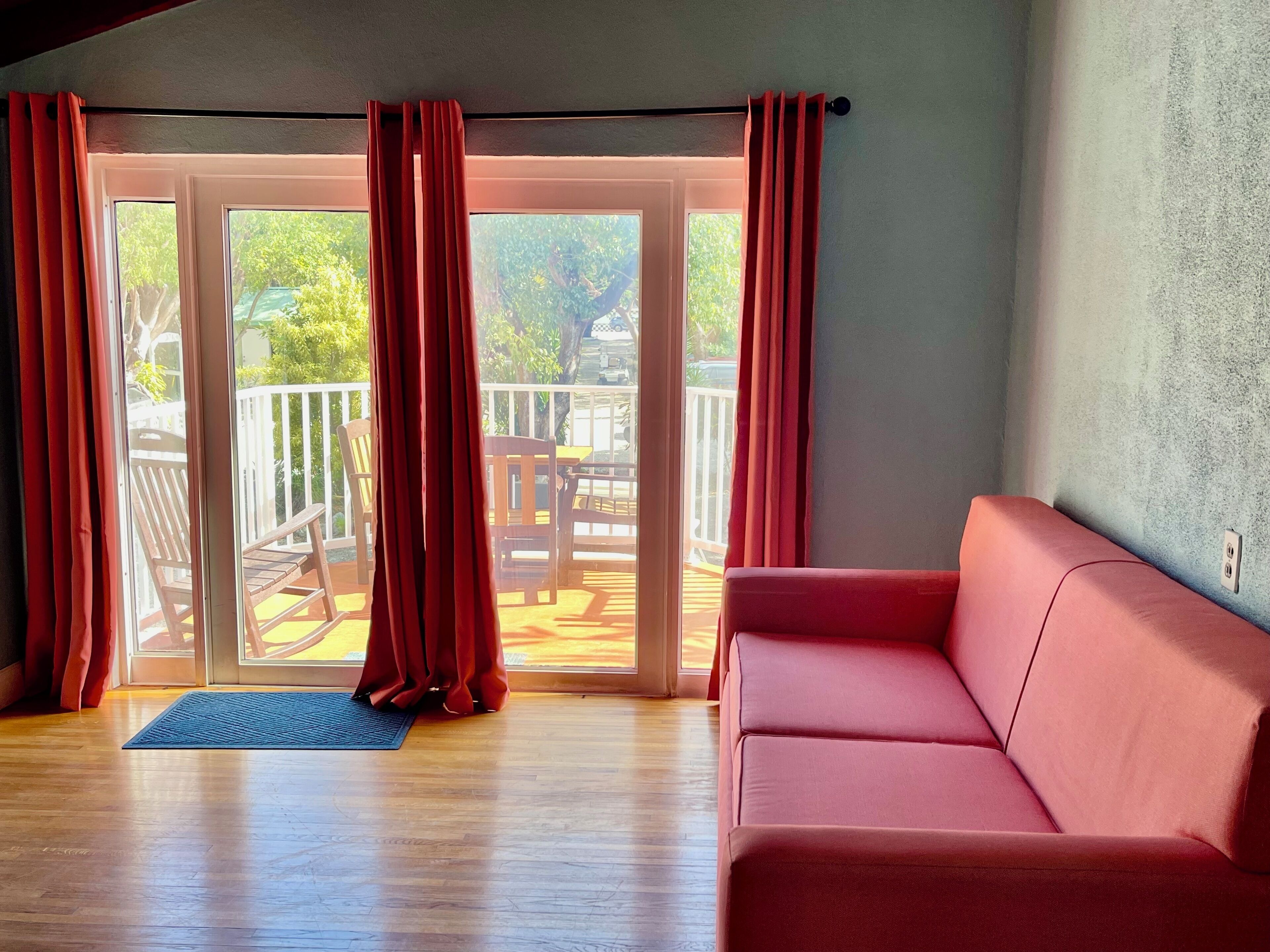 Et værelse med en rød sofa, trægulv og en skydedør med glas, der fører ud til en terrasse med træmøbler.