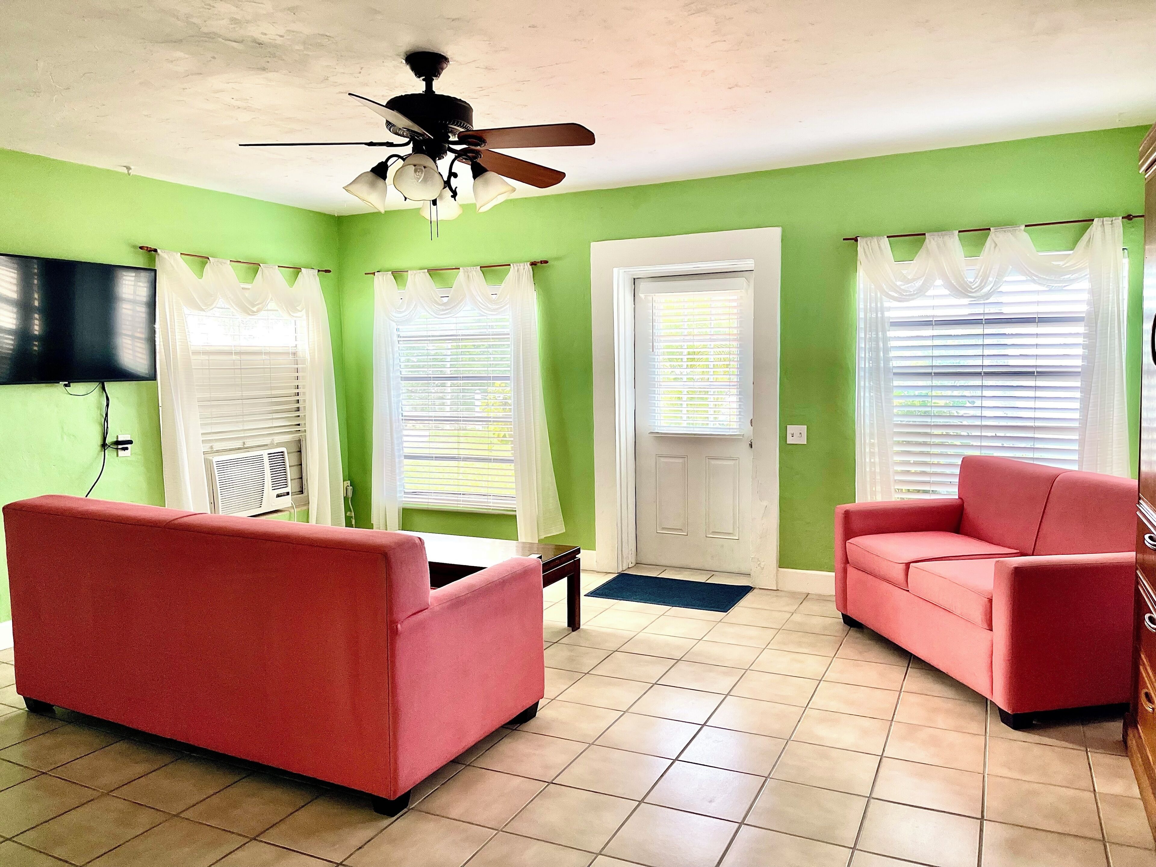 A living room with green walls, two red sofas, a ceiling fan, and a flat-screen TV mounted on the wall.