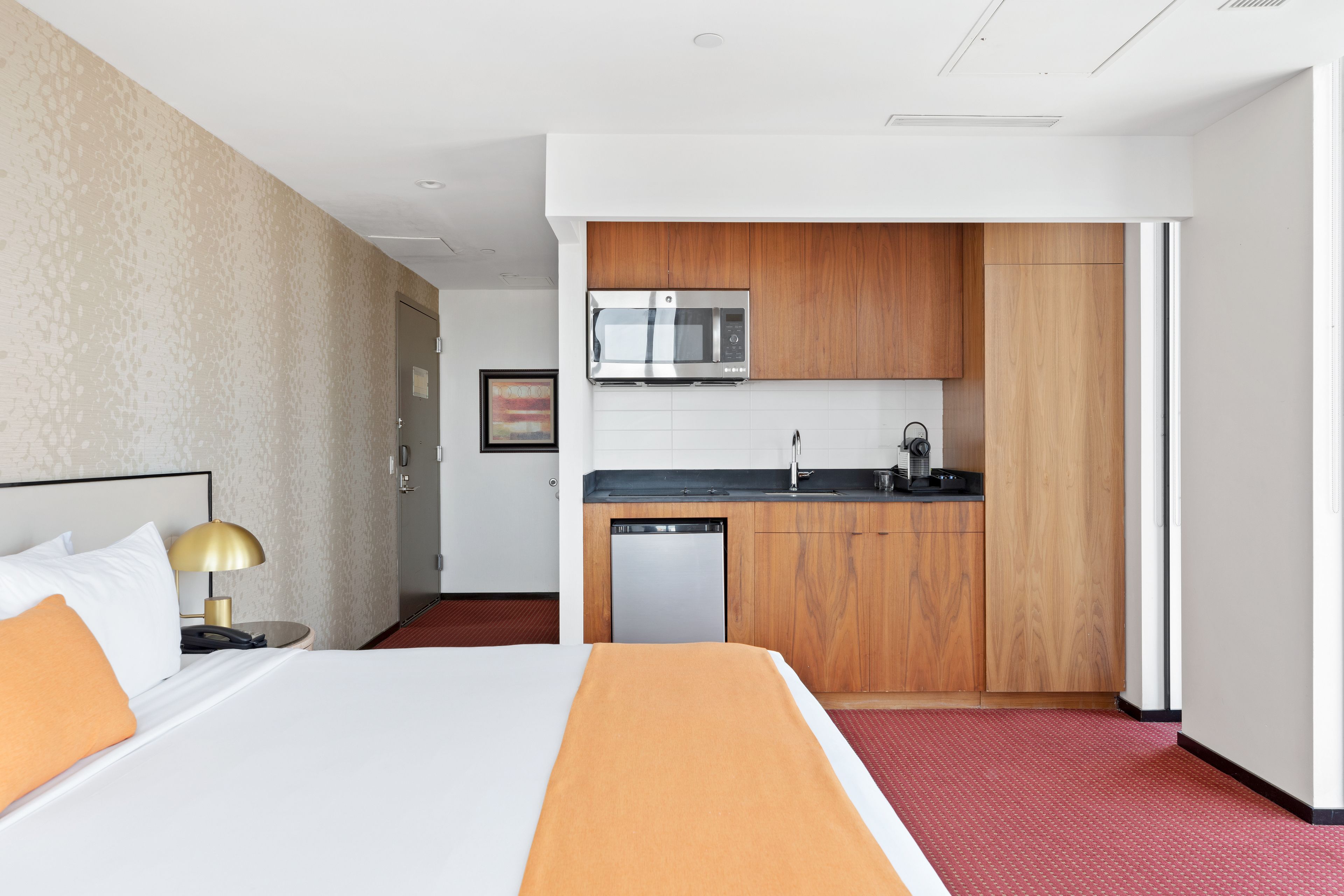Habitación de hotel con cama, mesita de noche con lámpara, kitchenette equipada con microondas, lavamanos y lavavajillas.