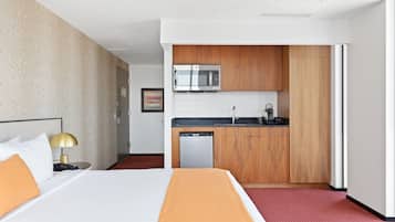 Habitación de hotel con cama, mesita de noche con lámpara, kitchenette equipada con microondas, lavamanos y lavavajillas.