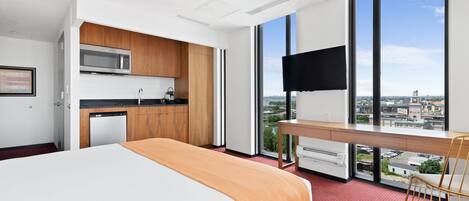 A modern hotel room with a bed, kitchenette, and a large window offering a city view.