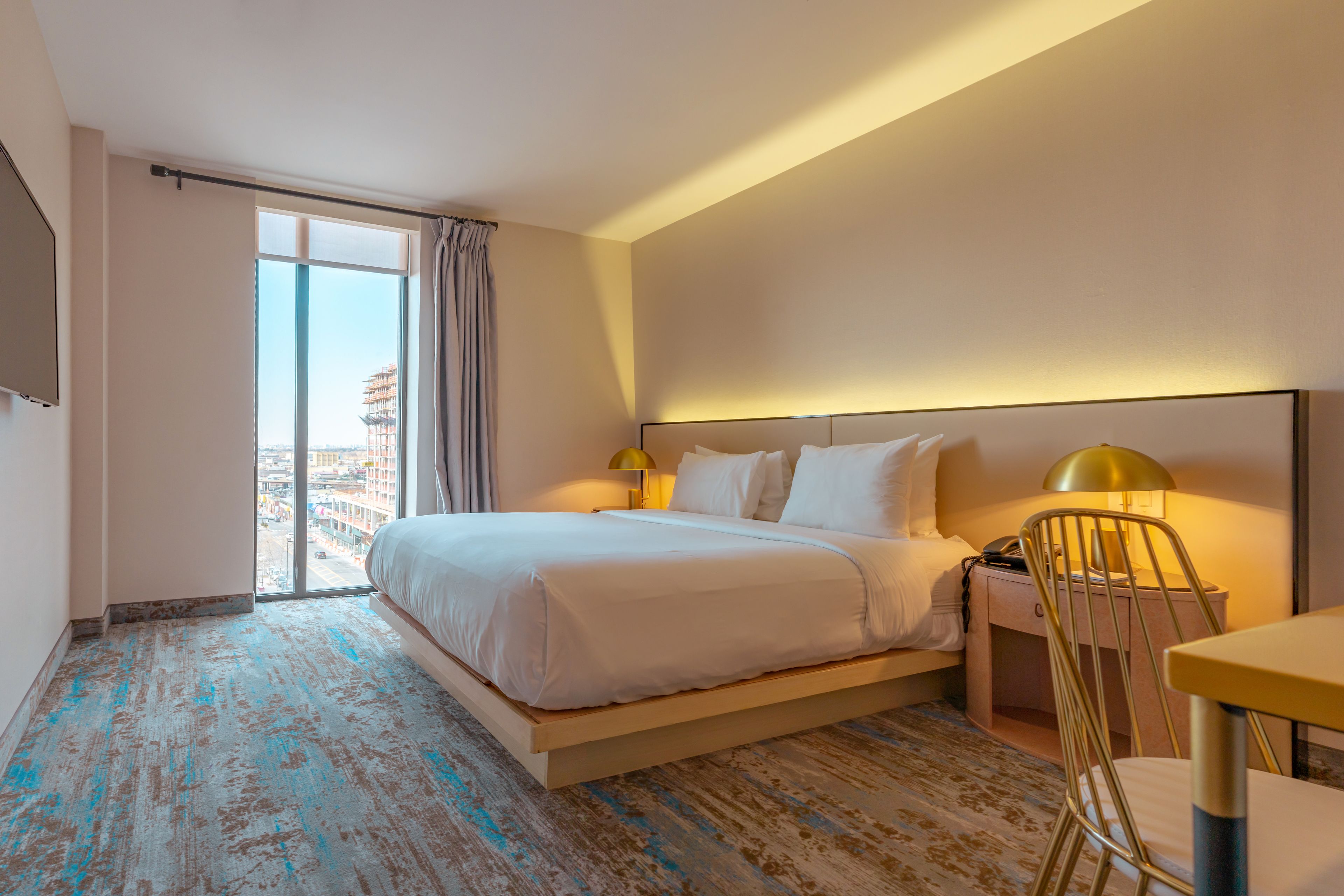 A hotel room with a large bed, a desk, a chair, a nightstand, and a window with a view of the city.