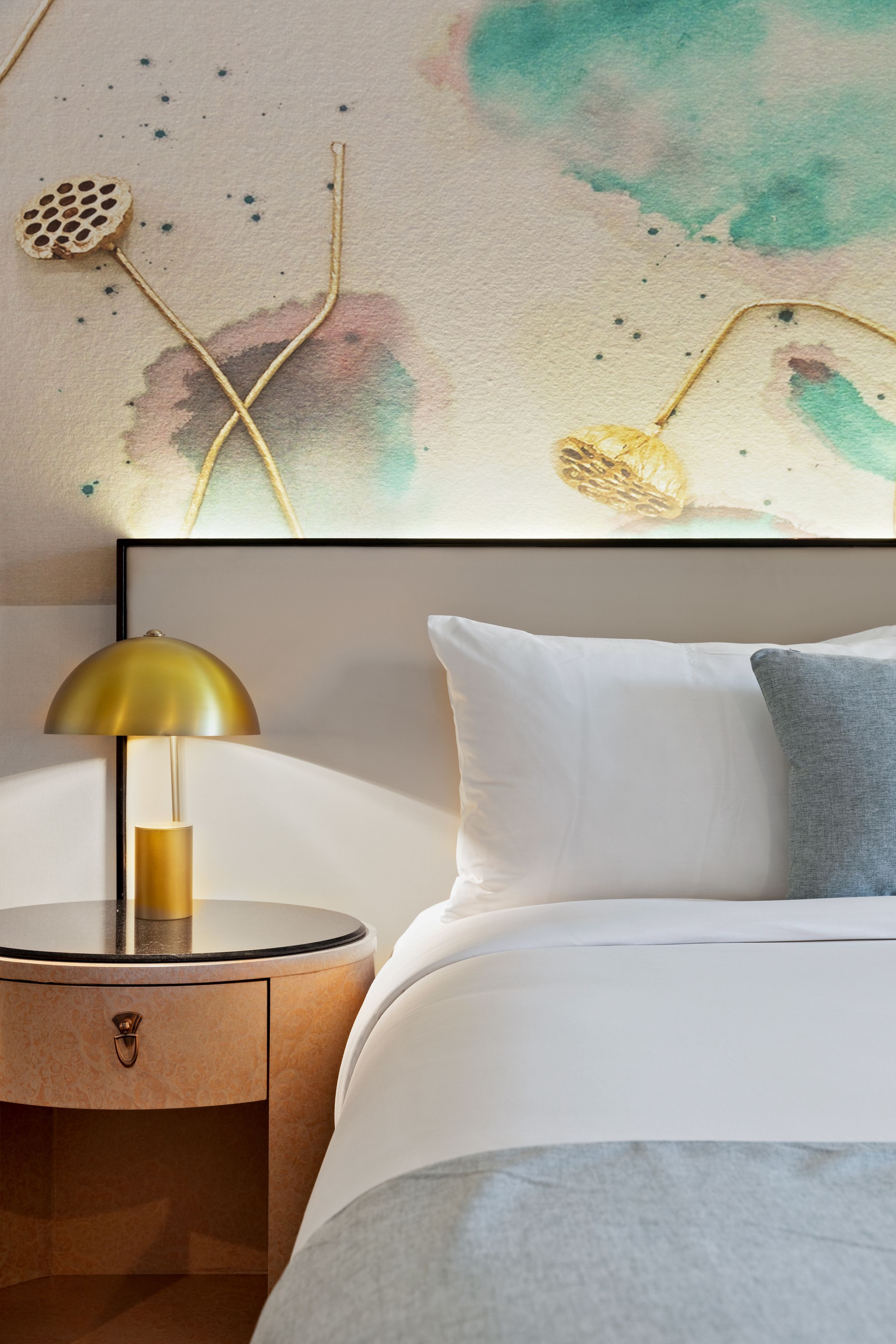 A neatly made bed with white and gray bedding, a gold lamp on a wooden bedside table, and a wall with abstract art.