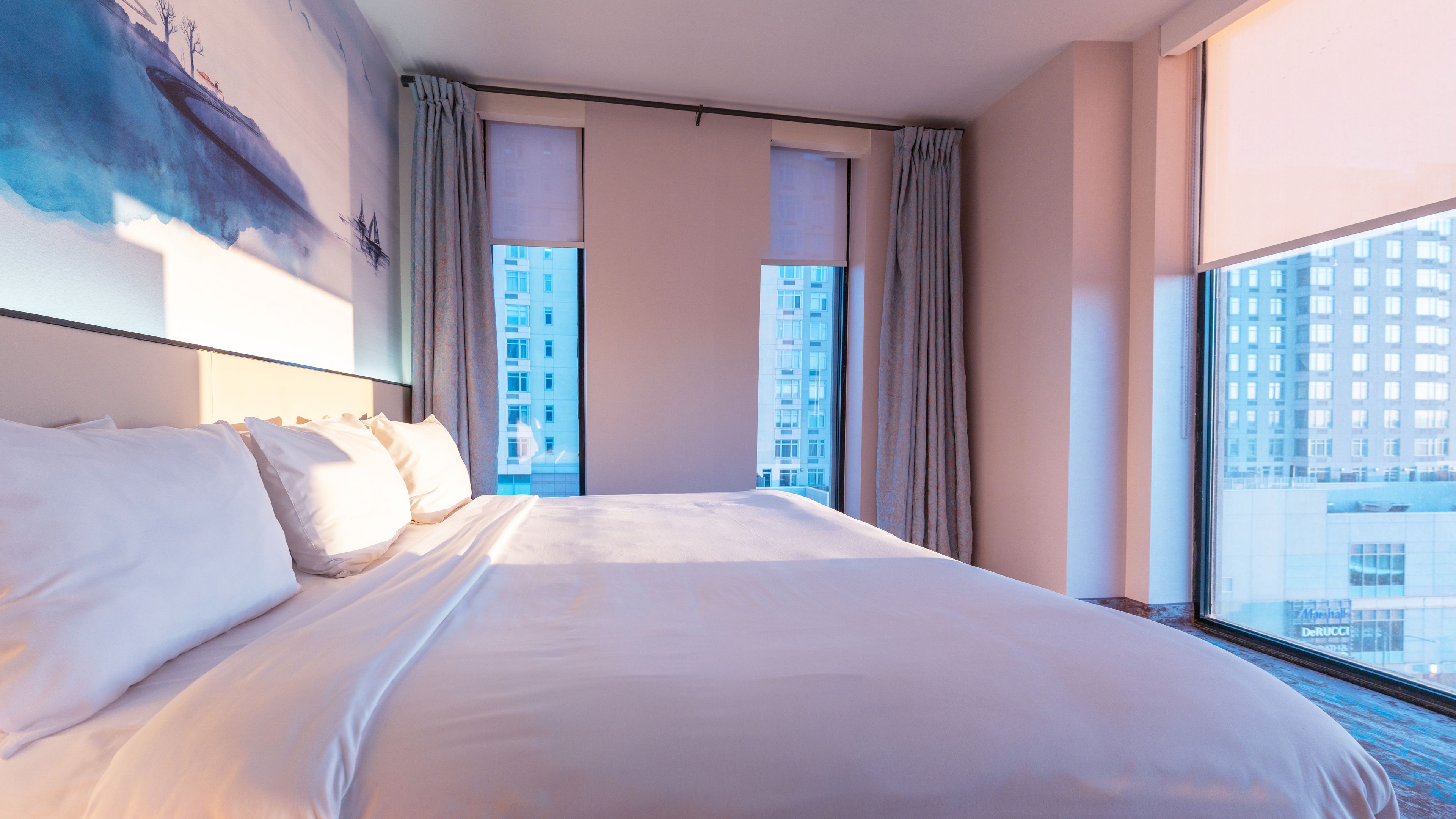 A hotel room with a large bed, a view of a cityscape through the window, and a painting on the wall.
