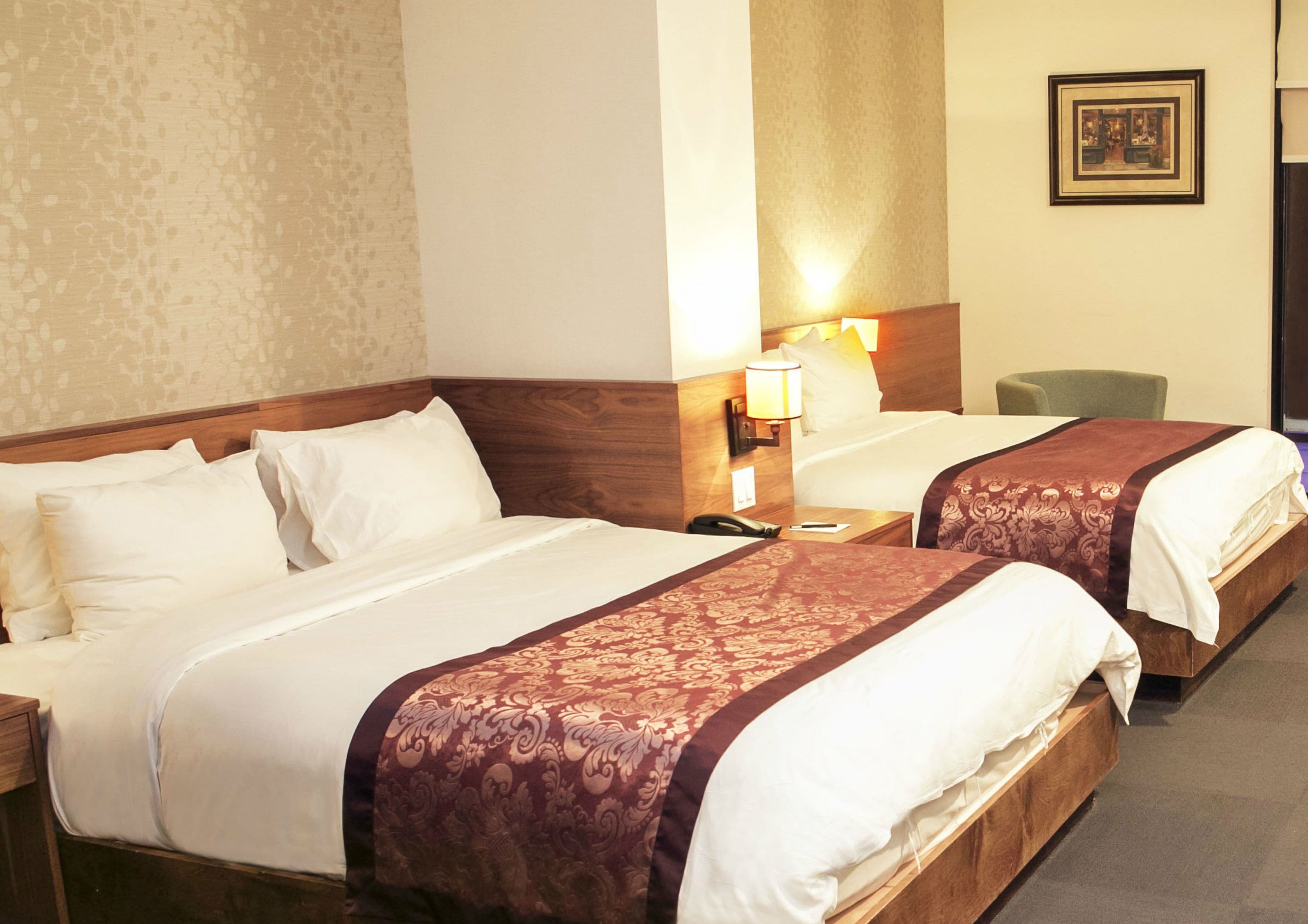 A hotel room with two beds, each with a wooden headboard and white bedding. There is a nightstand with a lamp and a phone on each bed. The room has a carpeted floor and a patterned wallpaper on the wall.