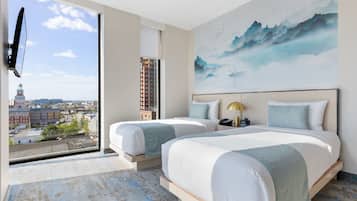 Ein Hotelzimmer mit zwei Betten, einem großen Fenster mit Blick auf die Stadt, einem Bild an der Wand und einem an der Wand befestigten Flachbildfernseher.