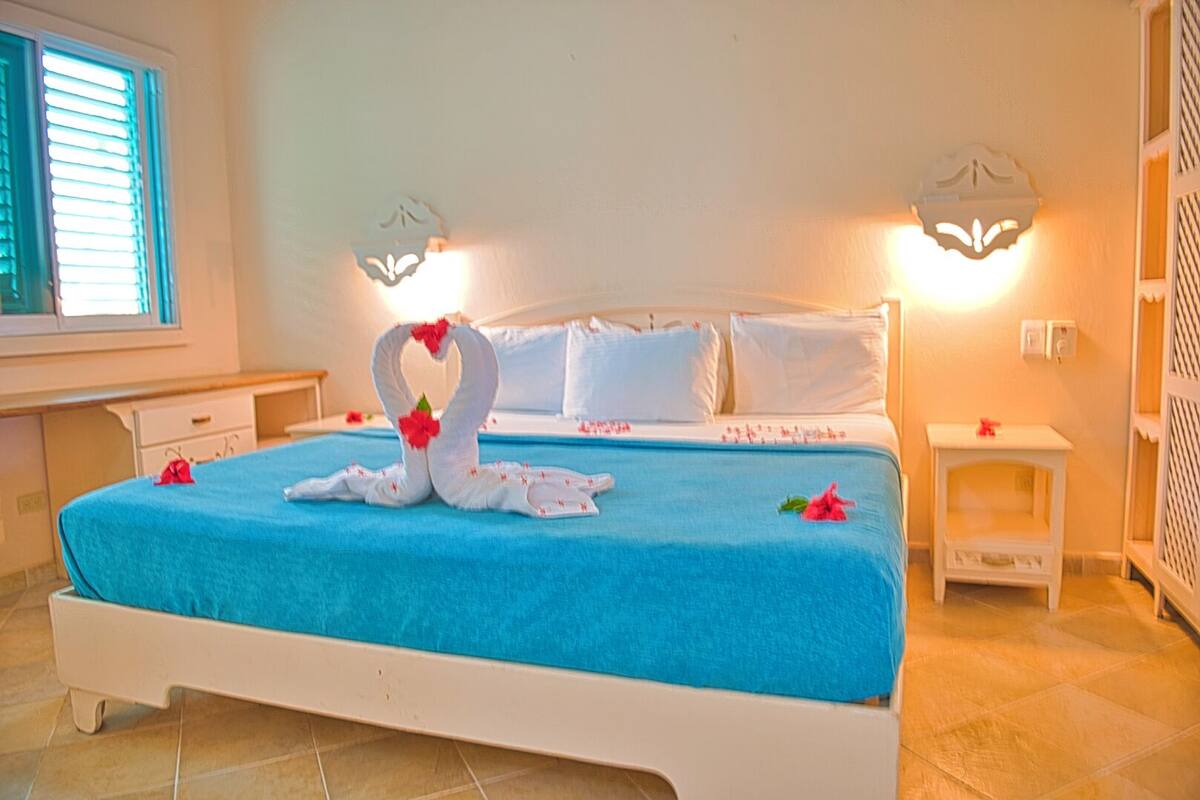 Una cama bien hecha con una colcha azul, dos toallas con forma de cisne y flores rojas. Hay dos mesitas de noche con lámparas, una ventana con persianas y una pared blanca con una lámpara decorativa.