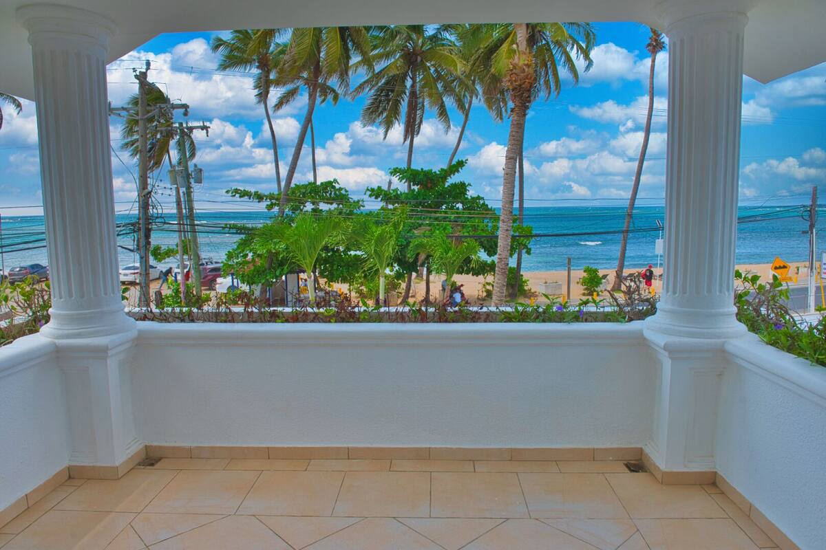 Un balcón con columnas blancas, piso de baldosas y vista a la playa, palmeras y el océano.