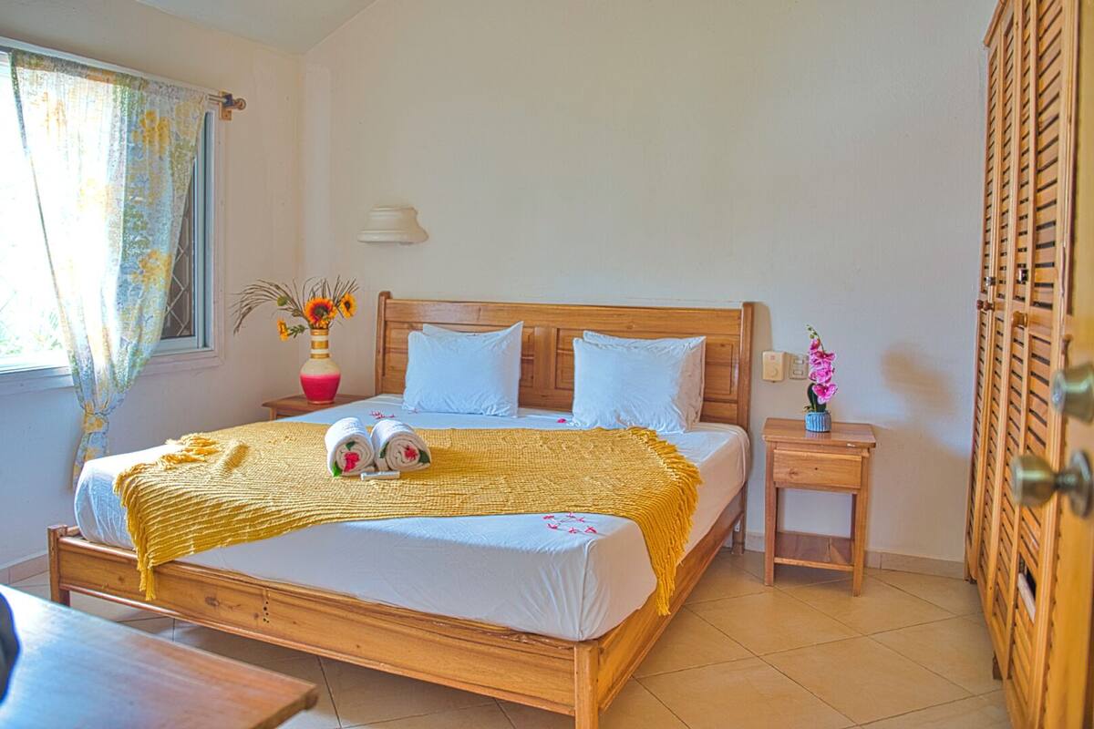 Una cama bien hecha con una manta amarilla, un cabecero de madera y dos almohadas. Se observa una mesita de noche con un jarrón de flores y una lámpara.