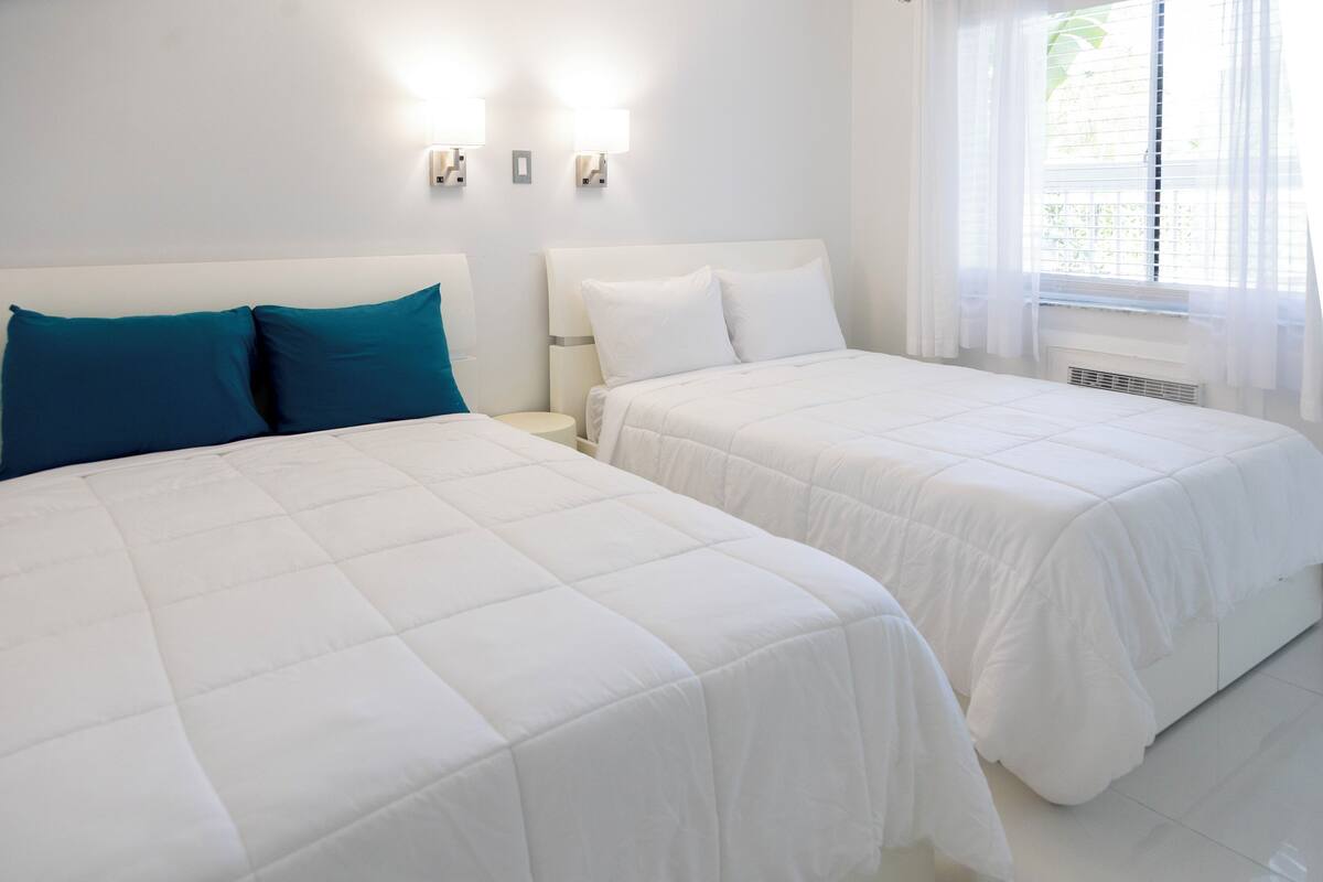 A hotel room with two beds, white bedding, a nightstand, wall-mounted lights, and a window with curtains.