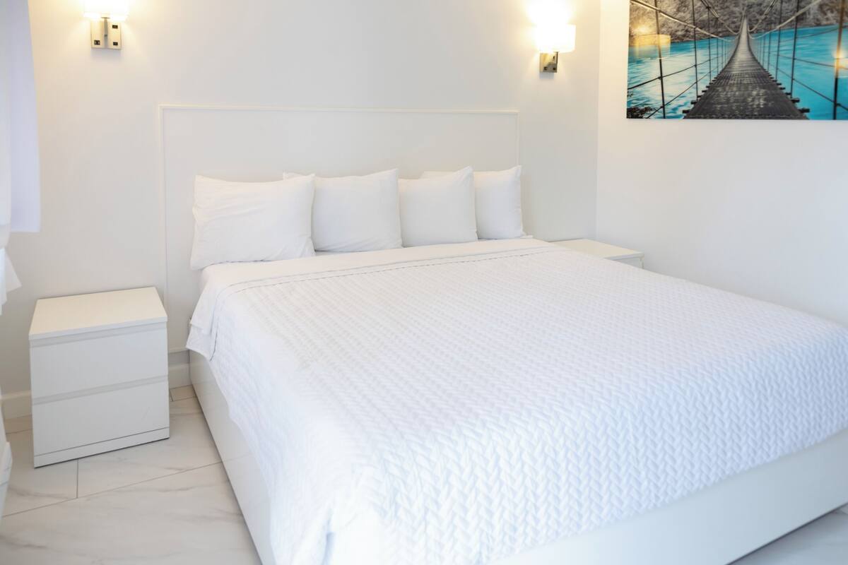 A white bed with a headboard, a bedside table, and a wall-mounted light fixture.