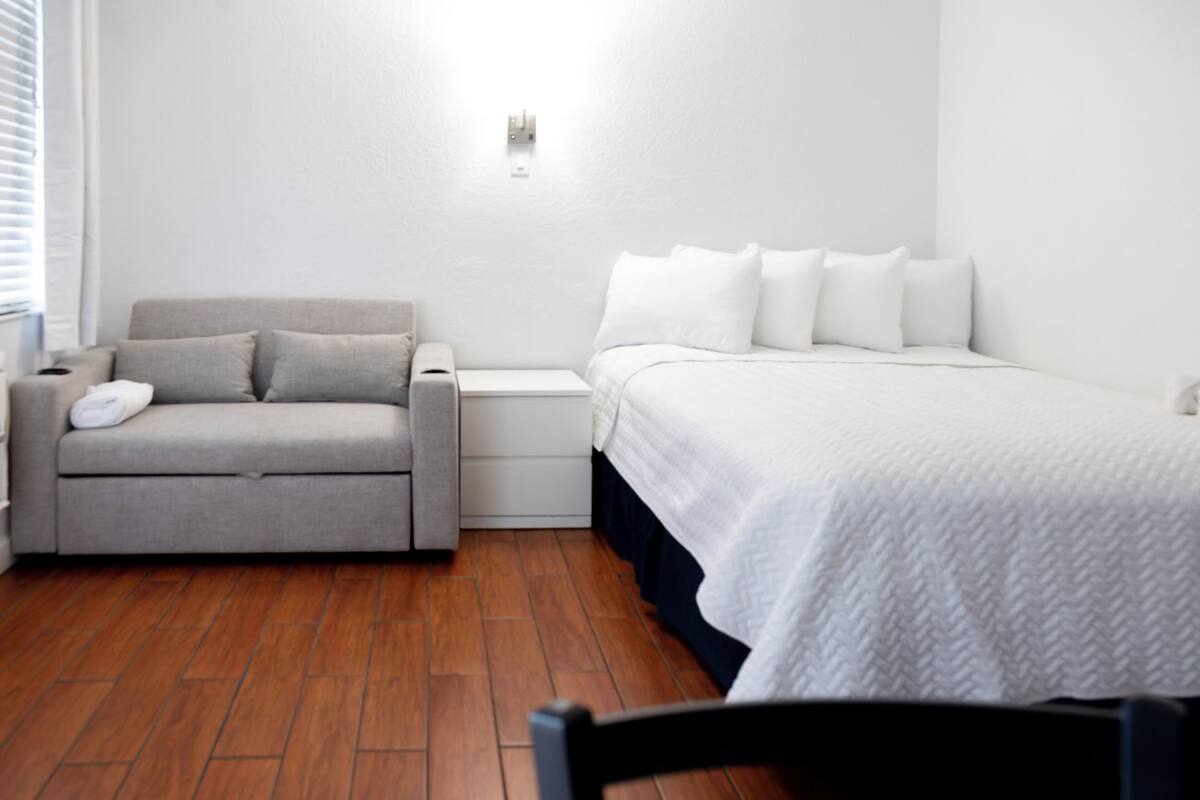 A bedroom with a bed, a grey sofa, a nightstand, and a wooden floor.