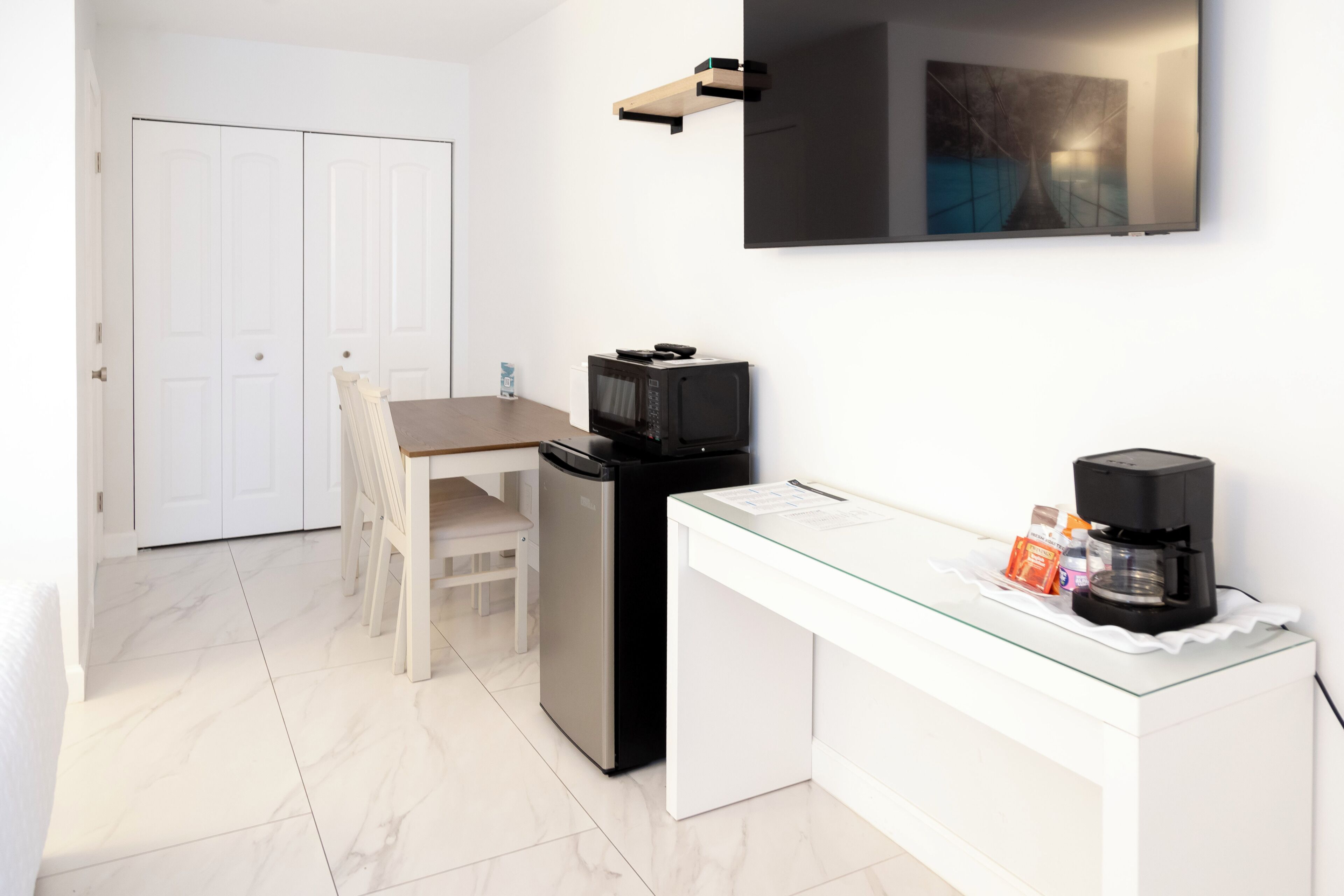 Una cocina compacta con microondas, refrigerador y cafetera sobre una mesa de vidrio.