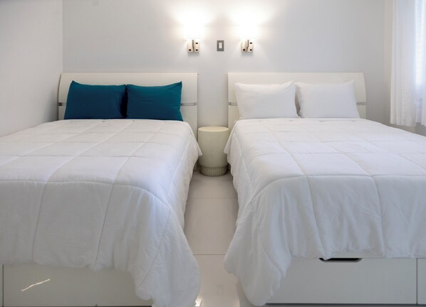 Une chambre d’hôtel avec deux lits, du linge de lit blanc, une table de chevet ronde et des lampes fixées au mur.
