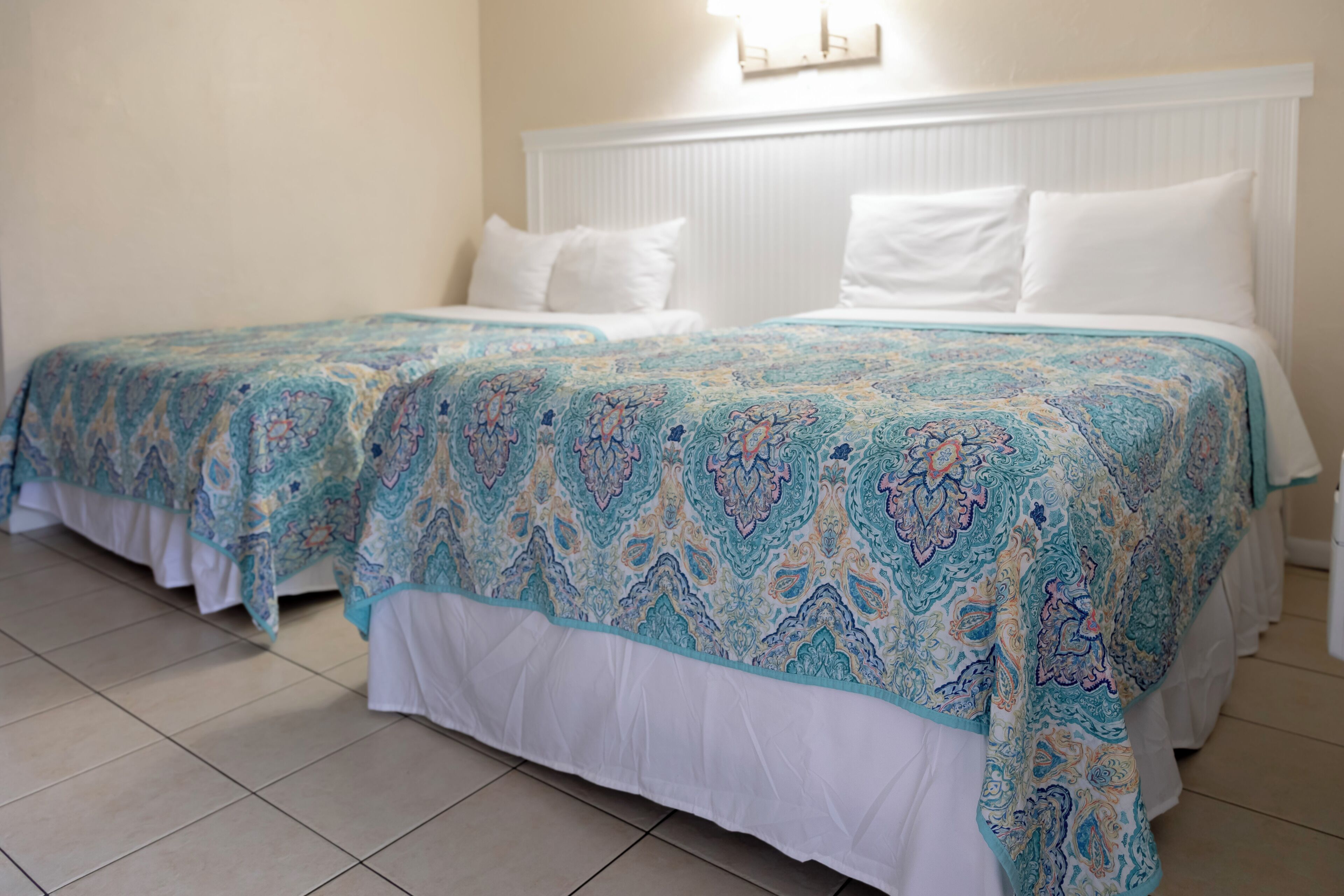 Un lit double avec une literie blanche et une couvre-lit à motifs, une tête de lit blanche et un luminaire fixé au mur.