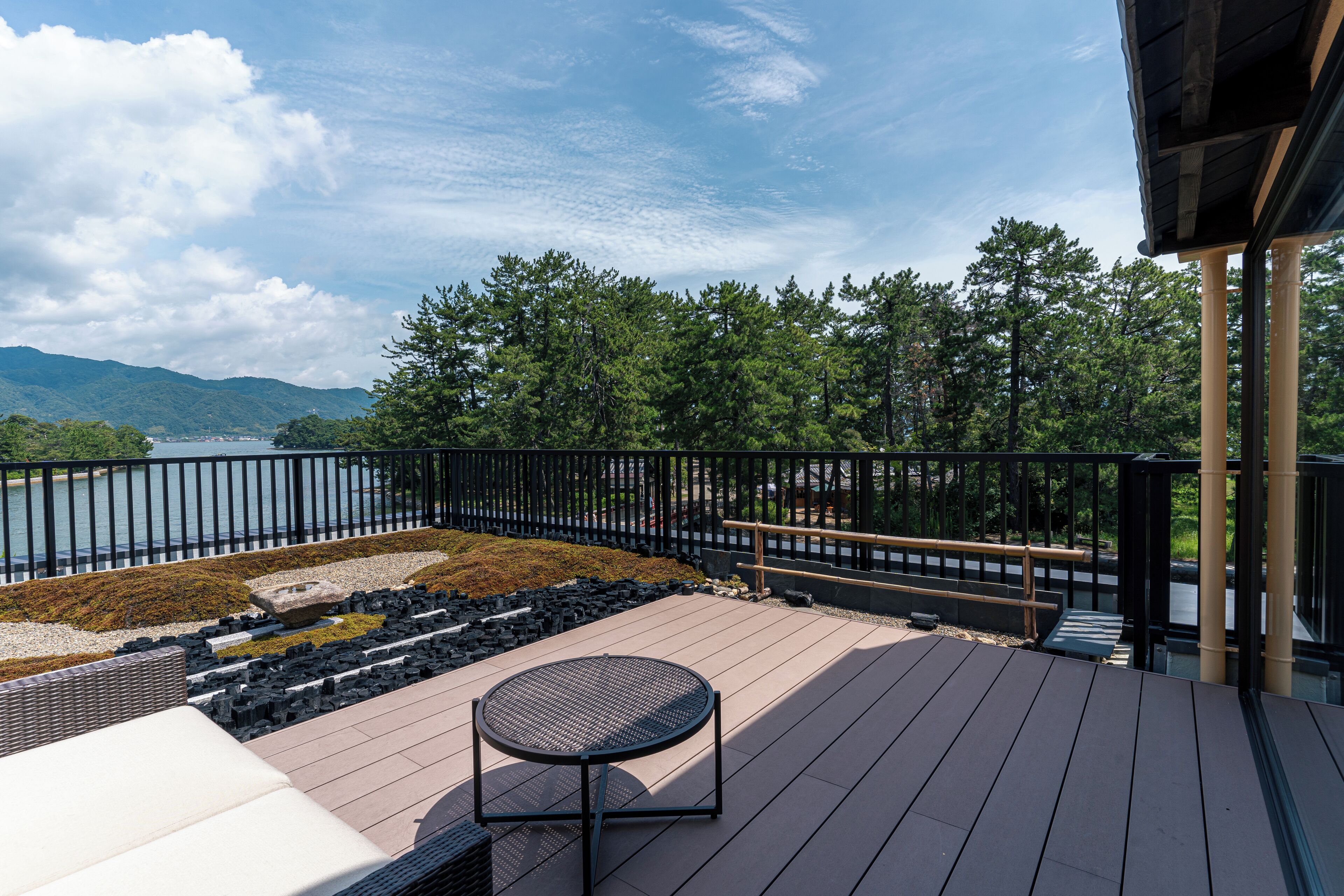 A deck with a round table and white sofa, overlooking a lake and forested hills.