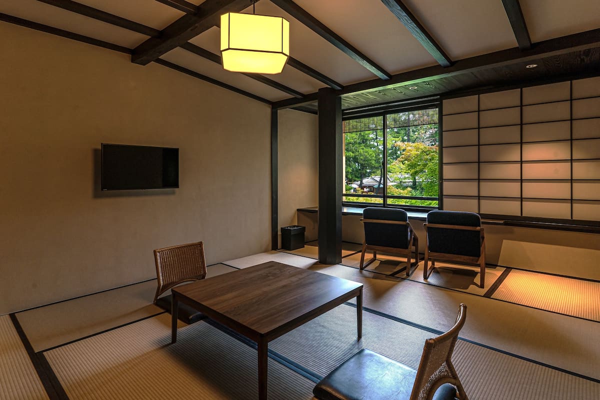 Ein traditioneller japanischer Raum mit Tatami-Böden, einem niedrigen Tisch, Stühlen, einer Hängeleuchte und Blick auf Grün durch ein Fenster.