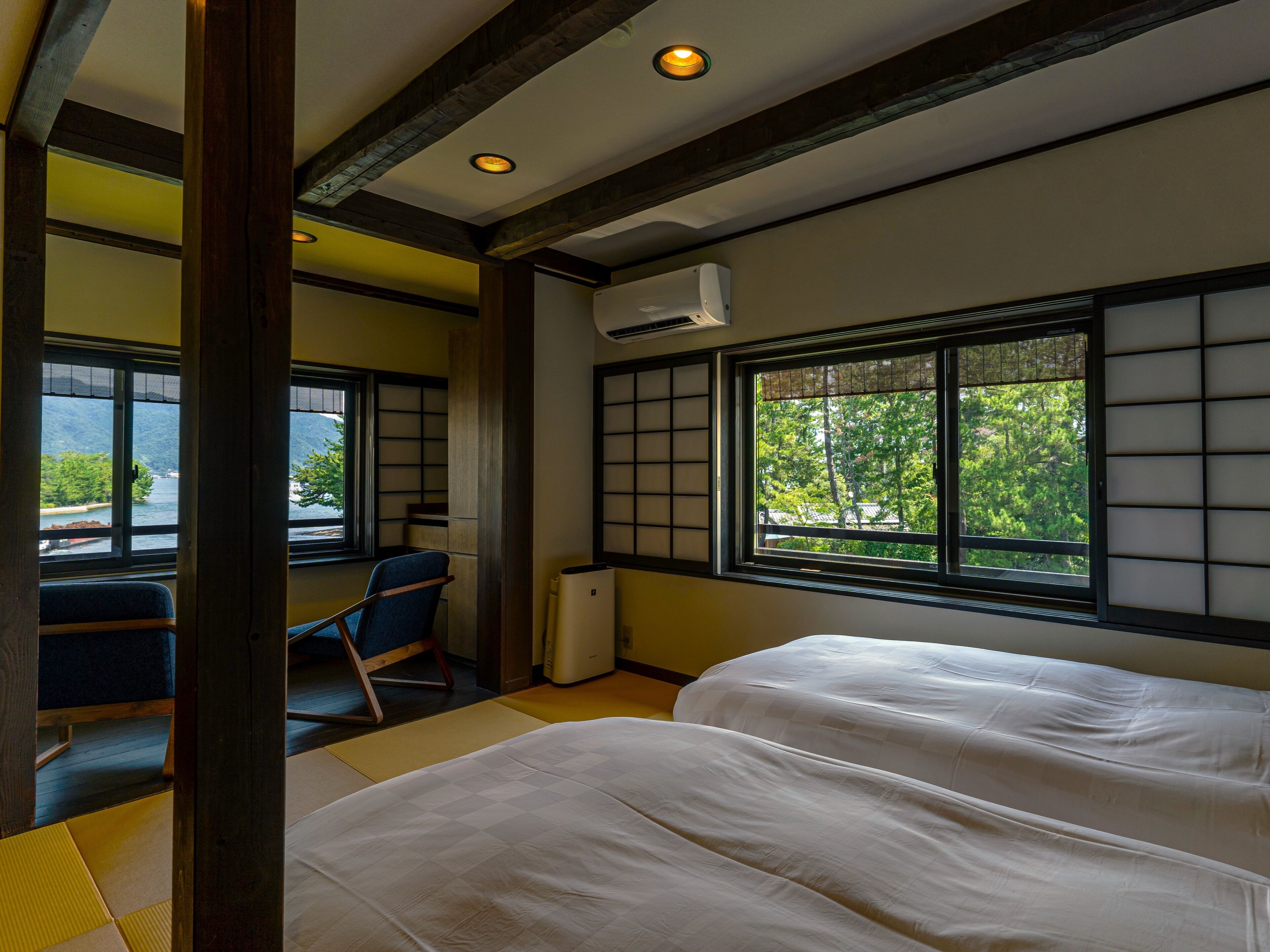 Ein traditioneller japanischer Raum mit Tatami-Boden, Futonbett, Schreibtisch mit Stuhl und Blick auf Grün durch ein Shoji-Fenster.